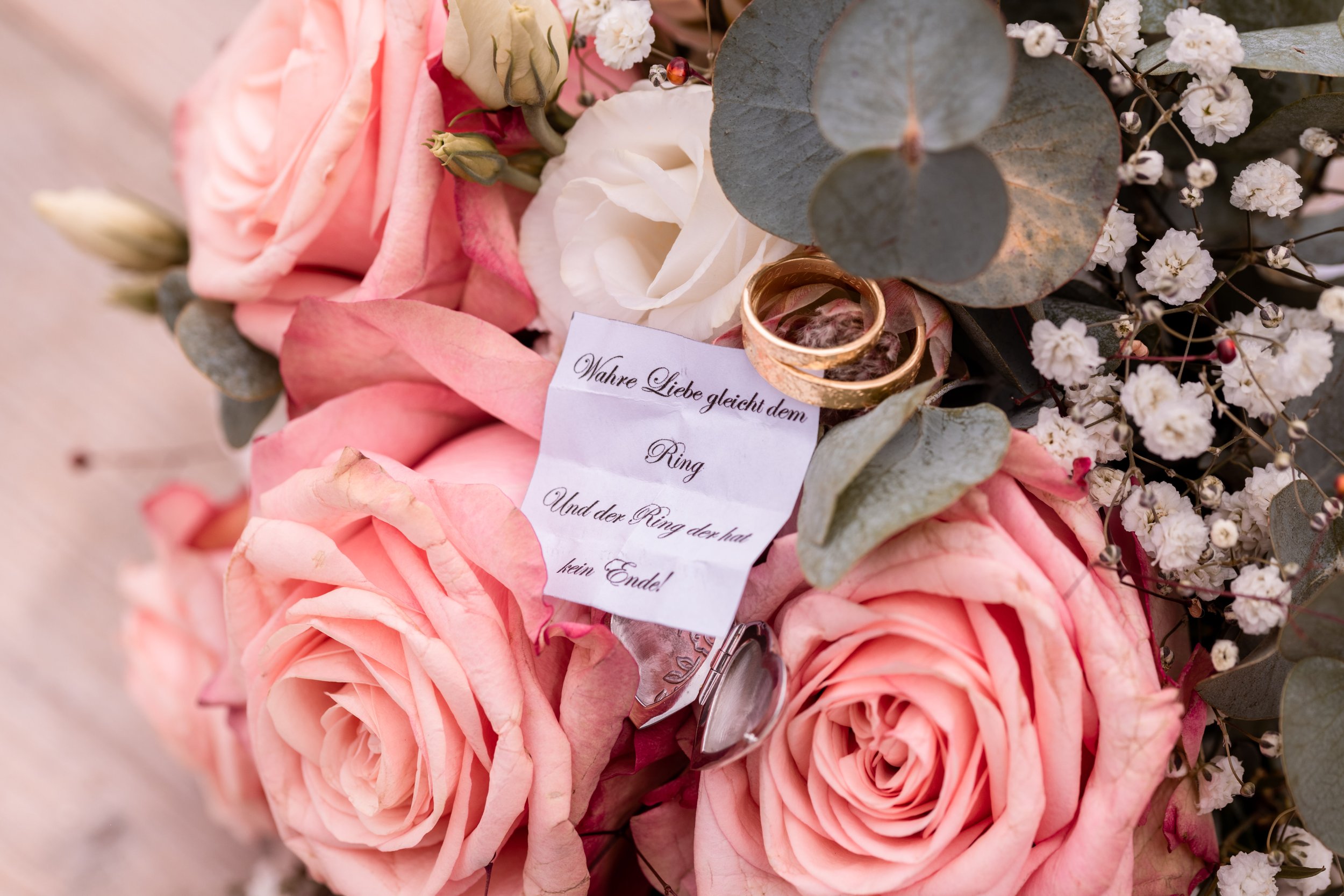 Ein Blumengesteck mit pinken Rosen, weißen Blumen und grünem Eukalyptus, auf dem zwei goldene Eheringe und eine kleine Notiz mit der Aufschrift "Wahre Liebe gleicht dem Ring. Und der Ring der hat kein Ende!" liegen.