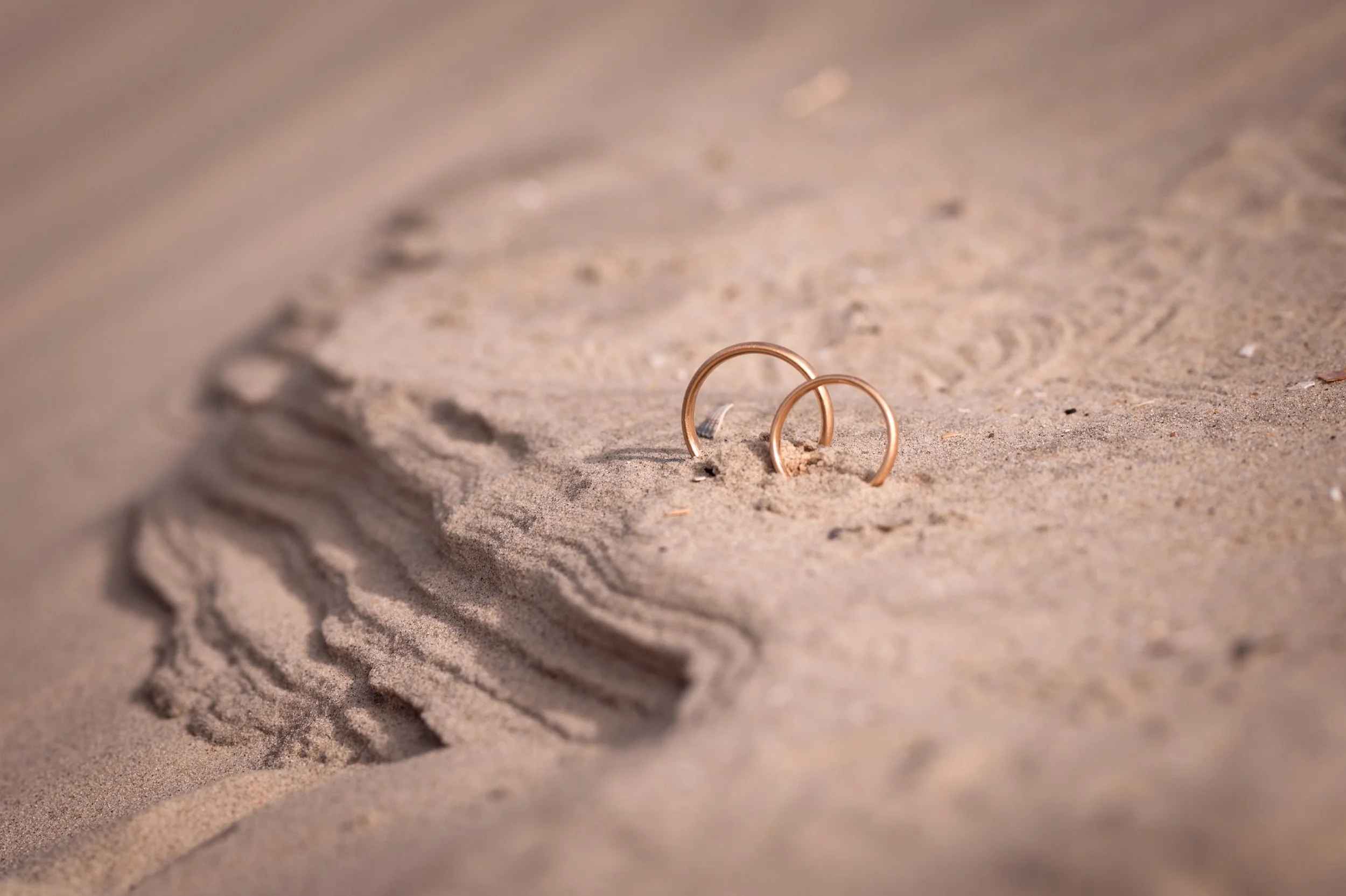 Zwei goldene Ringe im Sand am Strand, umgeben von Sandkörnern und kleinen Muscheln.
