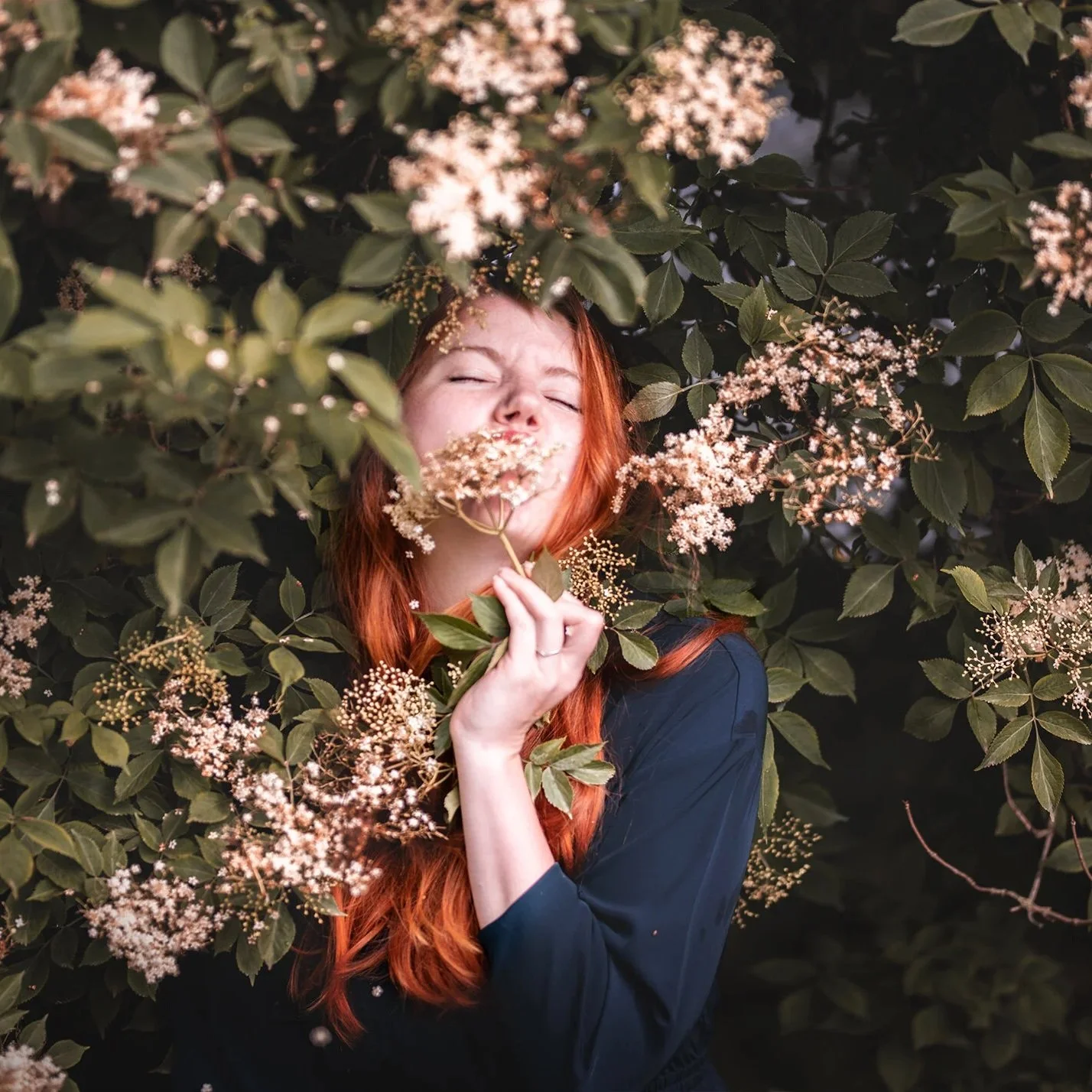 Eine rote-haired Frau umgeben von Blumen, hält eine Blume vor ihr Gesicht und scheint die Blume zu küssen oder zu schnuppern, mit ihren Augen geschlossen.