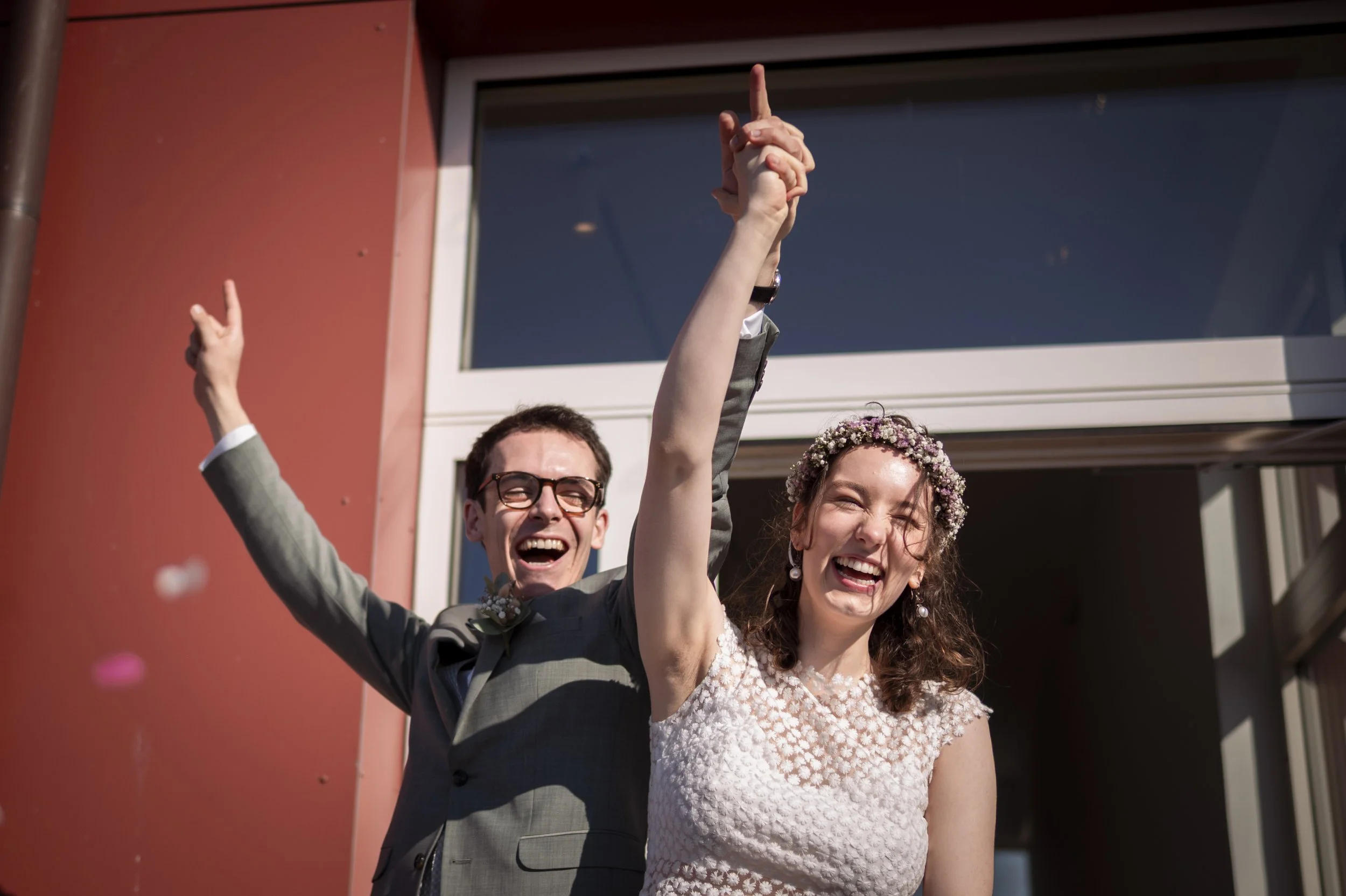 Ein glückliches Hochzeitspaar, lachend und in Freude, mit den Händen in der Luft, steht vor einem modernen Gebäude. Der Mann trägt einen Anzug und die Frau ein weißes, gewebtes Kleid mit Blumen im Haar.