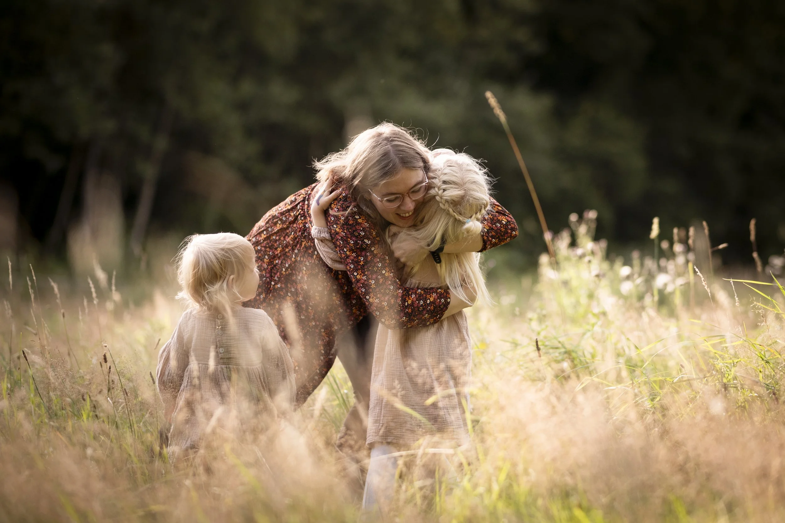 Familie beim Huggen in einem Wiesenfeld bei Sonnenuntergang, mit drei Mädchen und hüfthohen Gräsern.