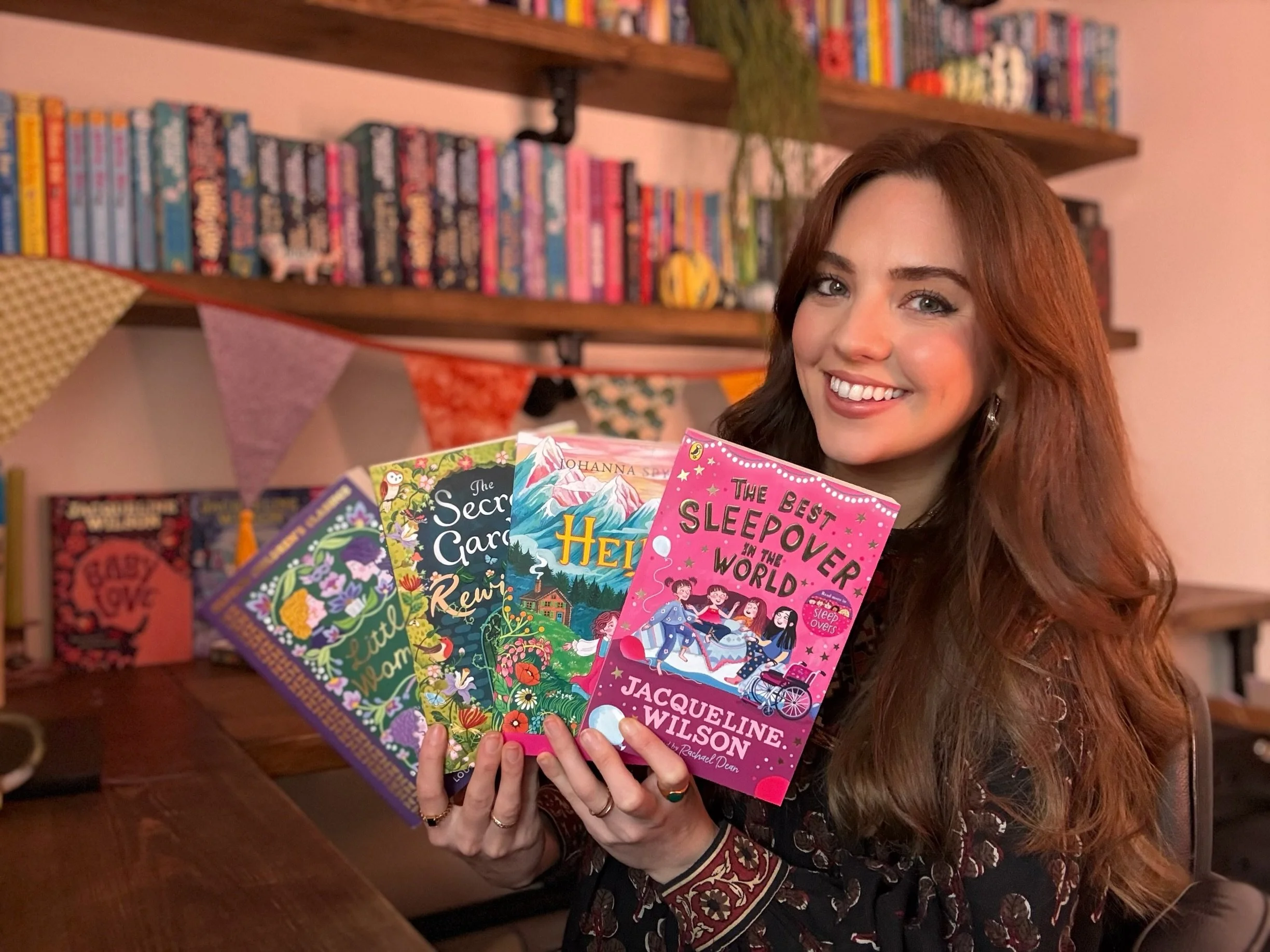A woman with long red hair and a patterned blouse holding several colorful books in front of a bookshelf decorated with bunting in a cozy room.