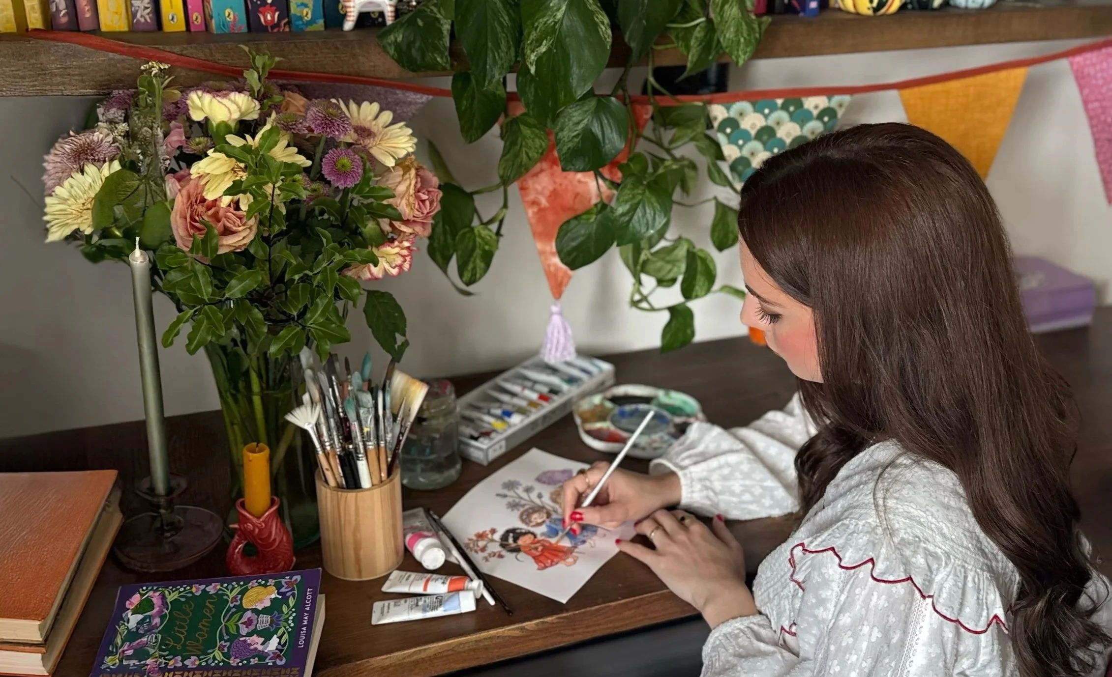 A young girl with long brown hair is painting a picture of two girls, one with black hair and one with red hair, while sitting at a wooden table. The table has art supplies including paintbrushes, paints, and coloring books. Behind her are books and a floral arrangement with pink, white, and purple flowers, and green leaves in a glass vase.