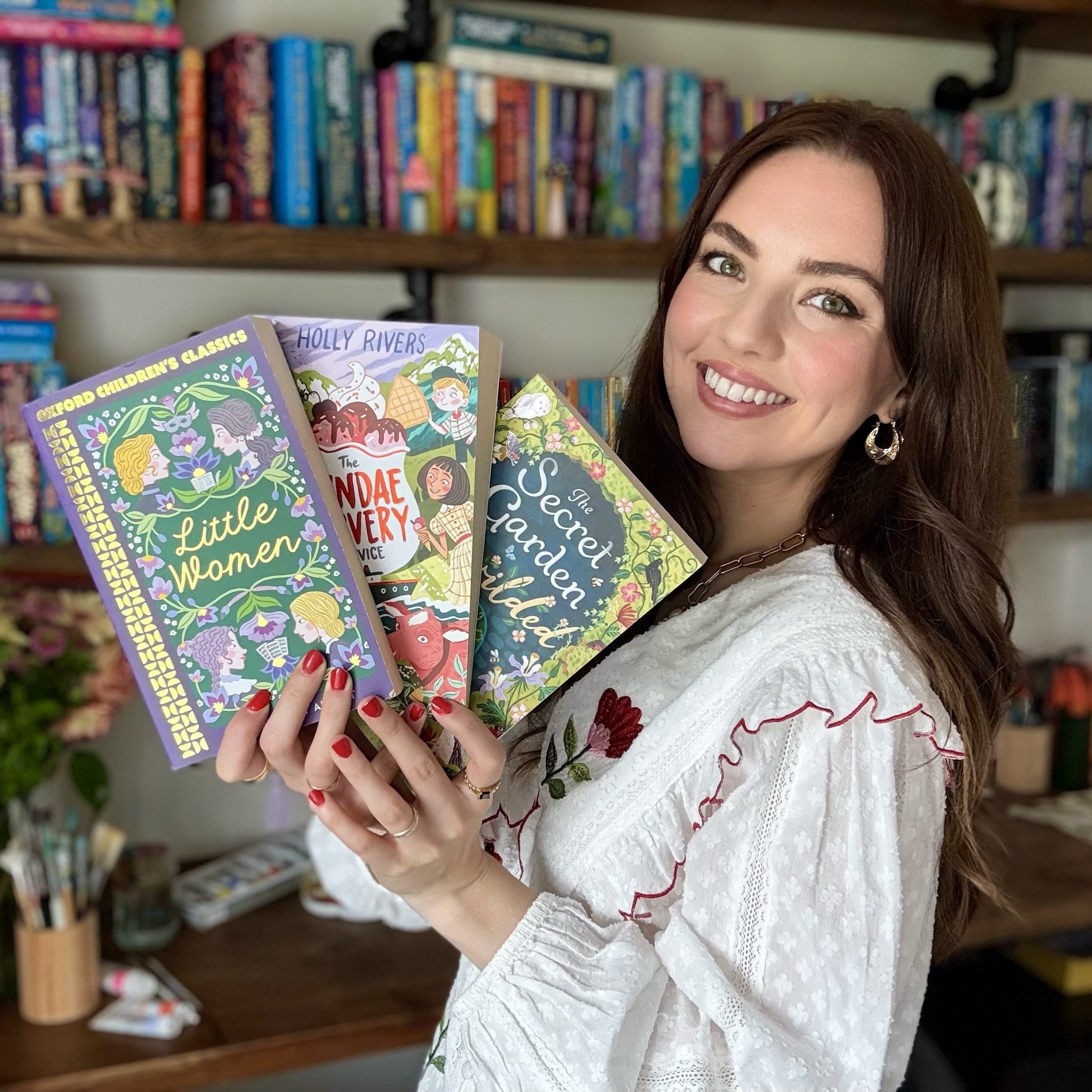 A woman with brown hair and earrings, smiling, holding a colorful book at a bookstore or library with shelves of books in the background.
