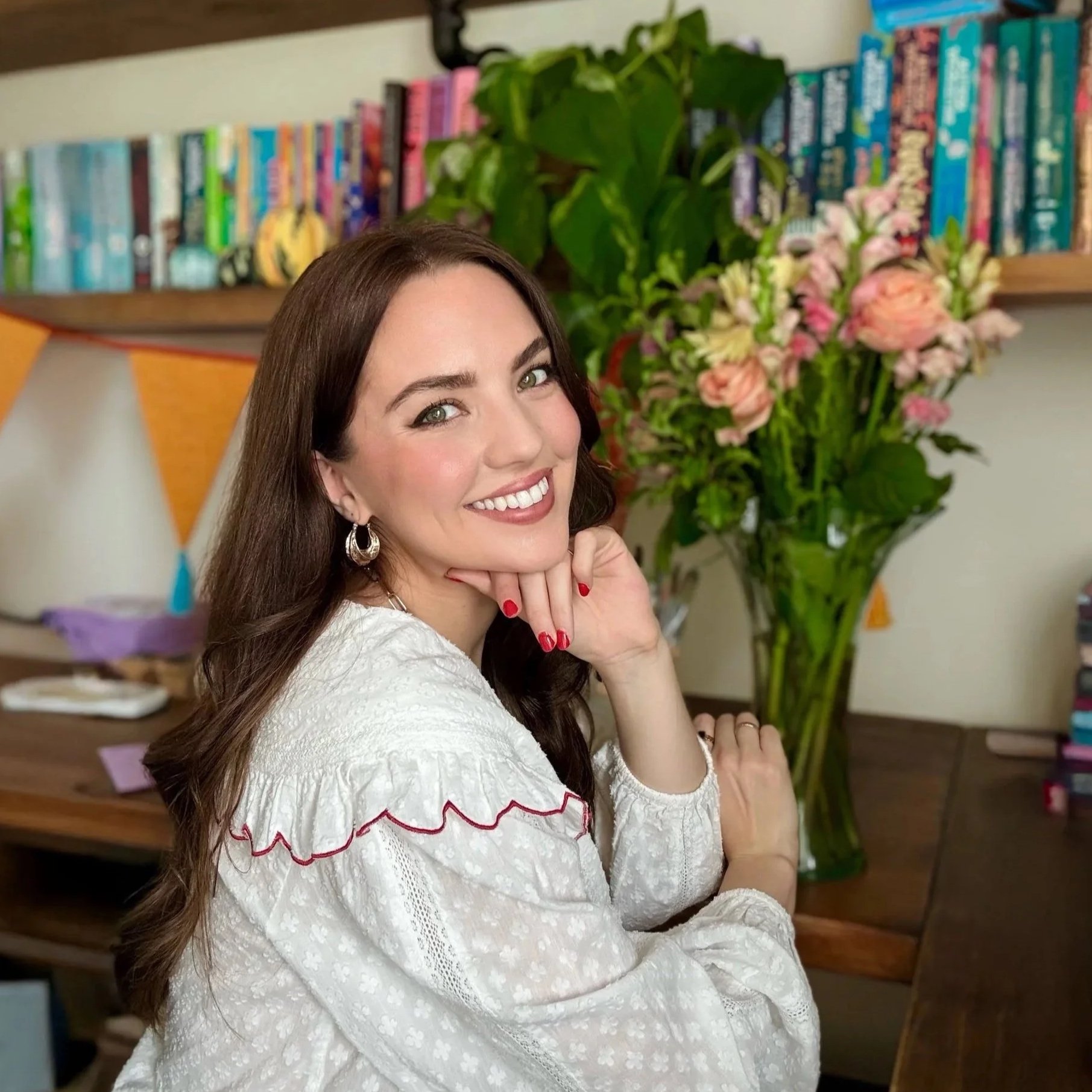 A woman with long brown hair and green eyes smiling. She is wearing a white blouse with red embroidery and large hoop earrings. In the background, there is a bookshelf with colorful books, a large green leafy plant, and a bouquet of pink and peach flowers.
