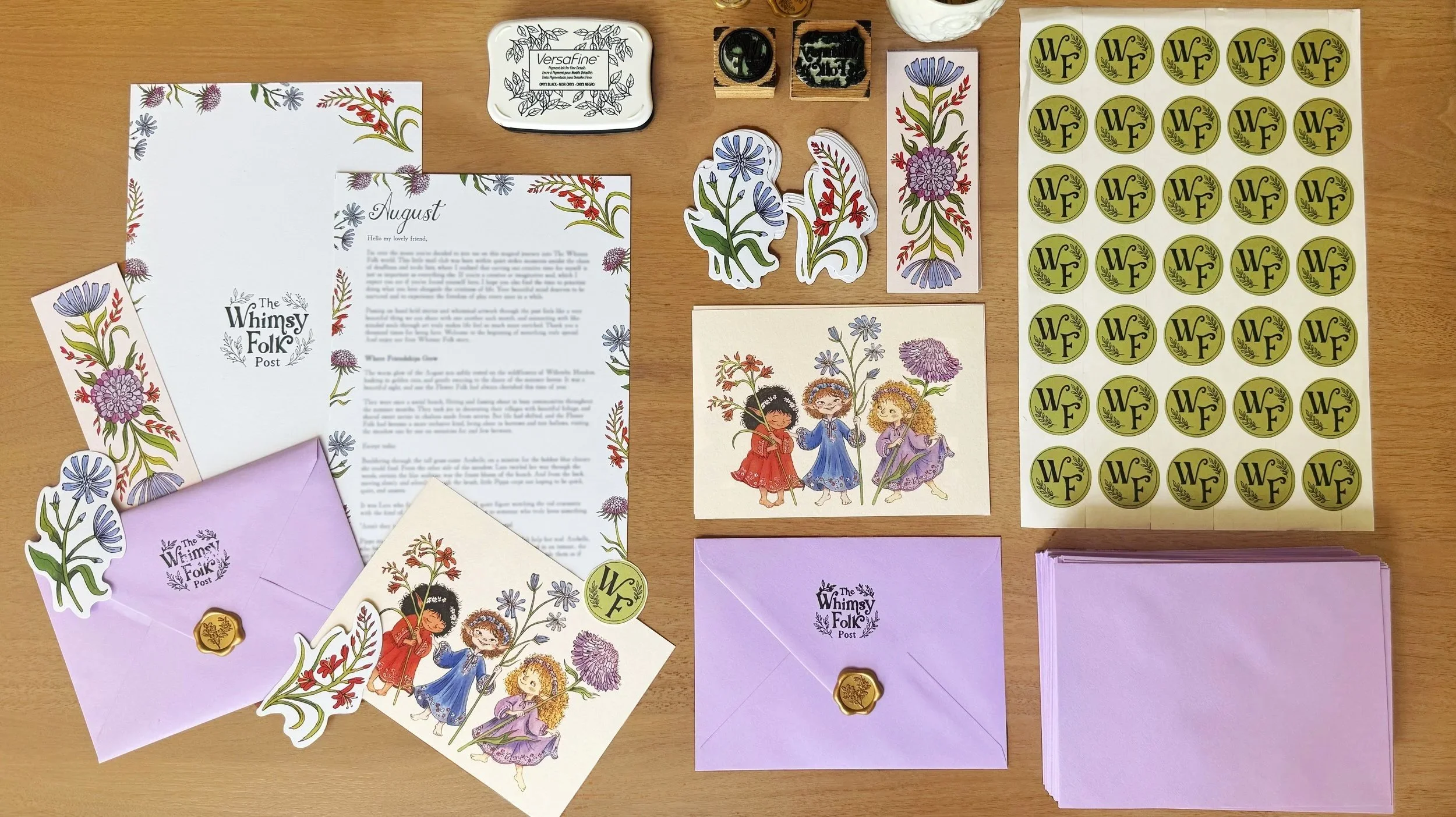 Flat lay of greeting cards, stickers, envelopes, and stamps with floral and folk art designs, including children holding flowers, purple and blue flowers, and decorative letter stickers on a wooden surface.