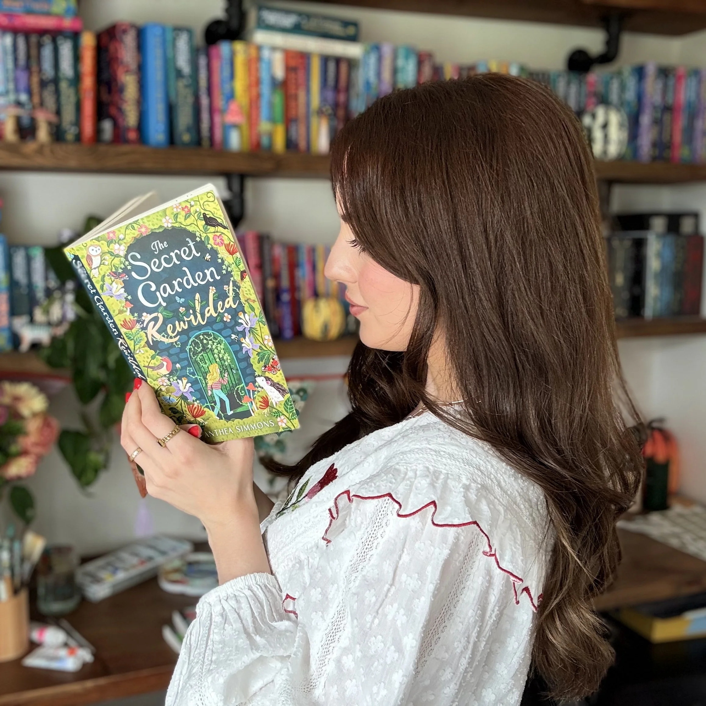A woman with brown hair reading a book titled "The Secret Garden Rewilded" in a library or bookstore, with shelves of colorful books in the background.