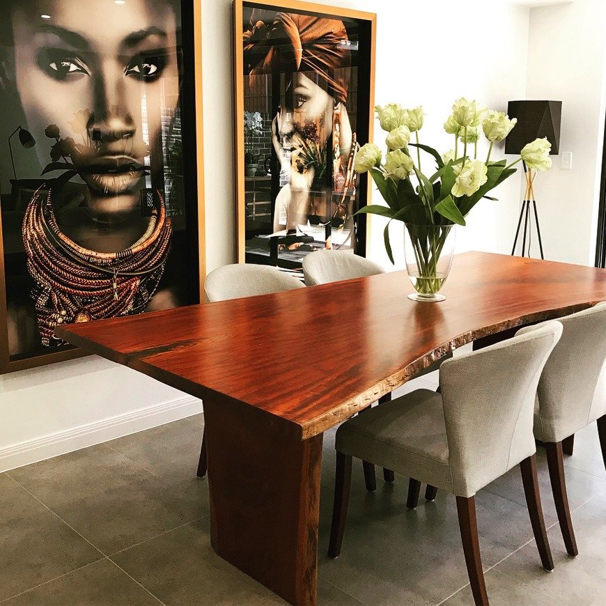Interior designed living room featuring a large wooden table, six upholstered chairs, large portrait paintings on the wall, vase with white flowers, and a floor lamp.