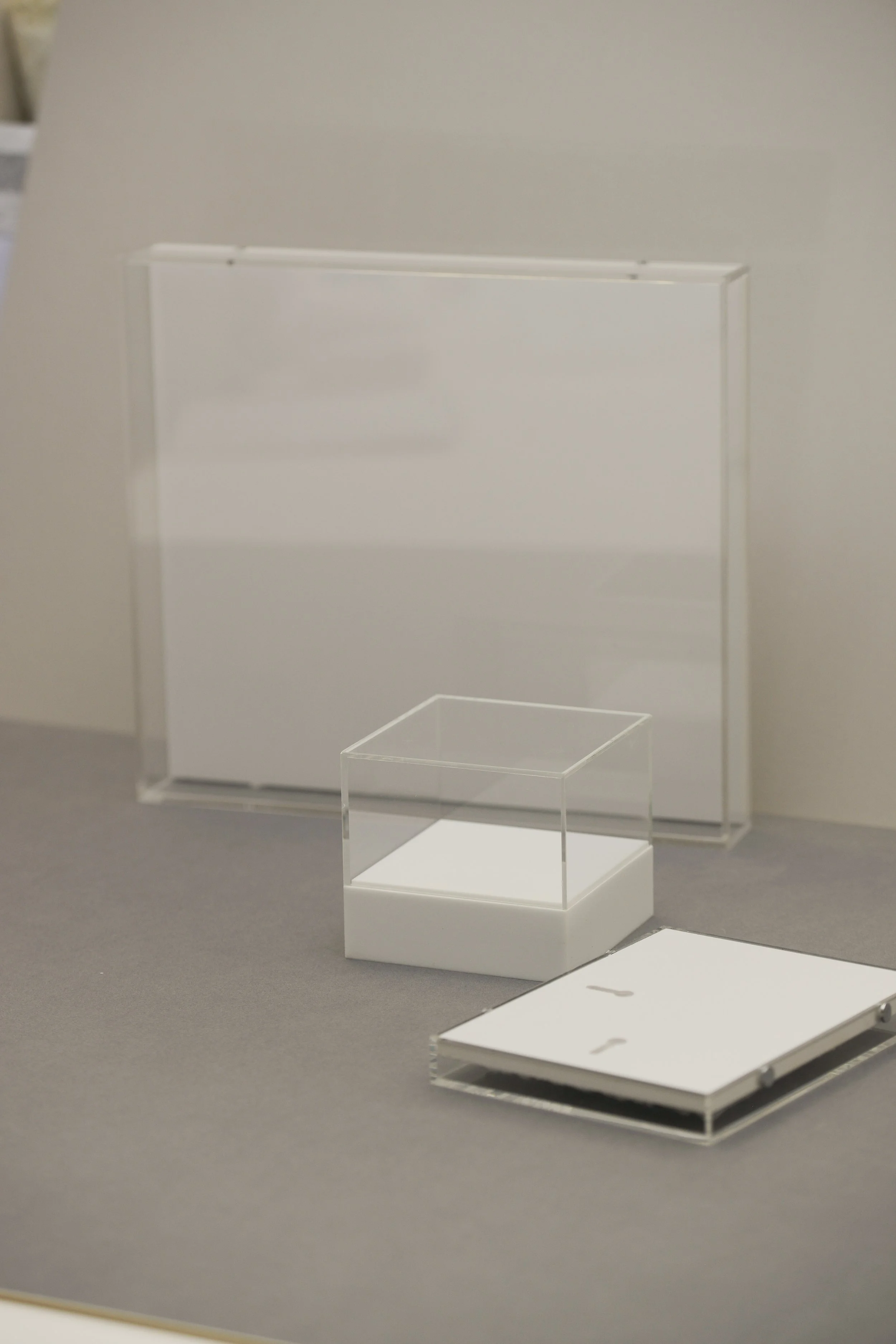 A clear plastic display case with a white base, a larger white panel or board behind it, and a small stack of white trays or panels on a gray surface.