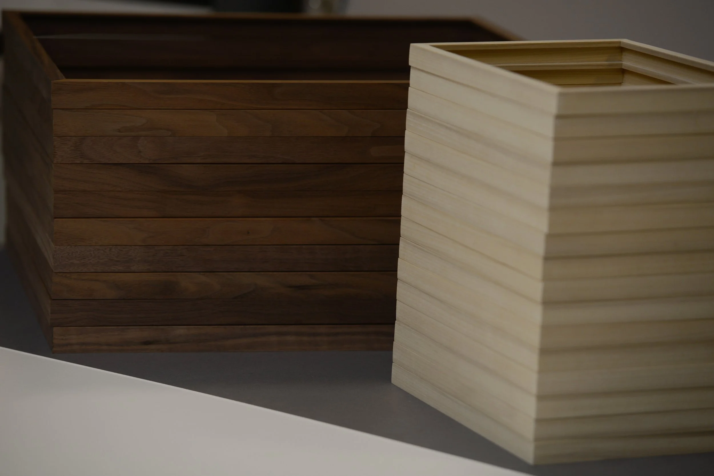 A close-up of two rectangular wooden boxes, one darker stained wood and one light natural wood, placed on a grey surface with a white background.