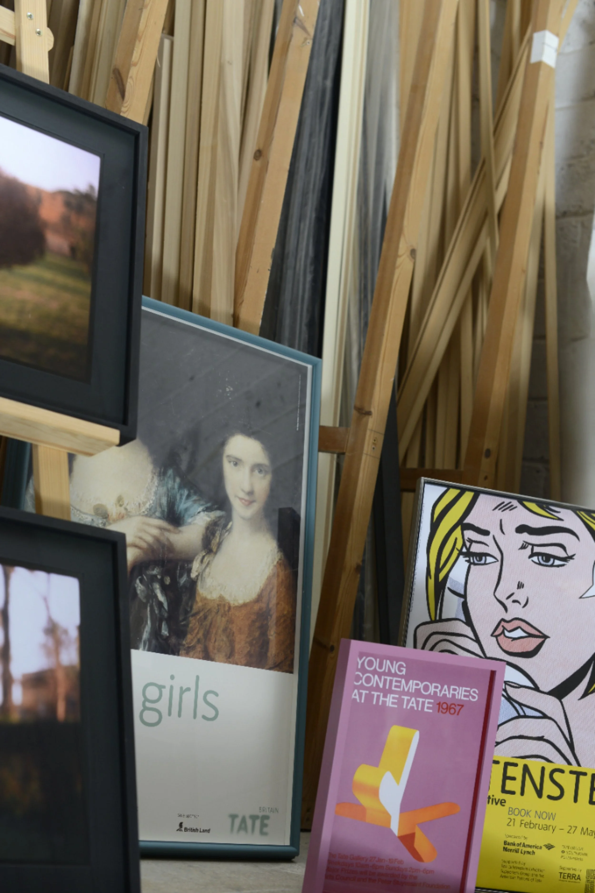 Collection of framed posters and artworks leaning against a wall with wooden planks in the background. The posters include an image of the Mona Lisa, a comic-style face, and promotional materials for art exhibitions.