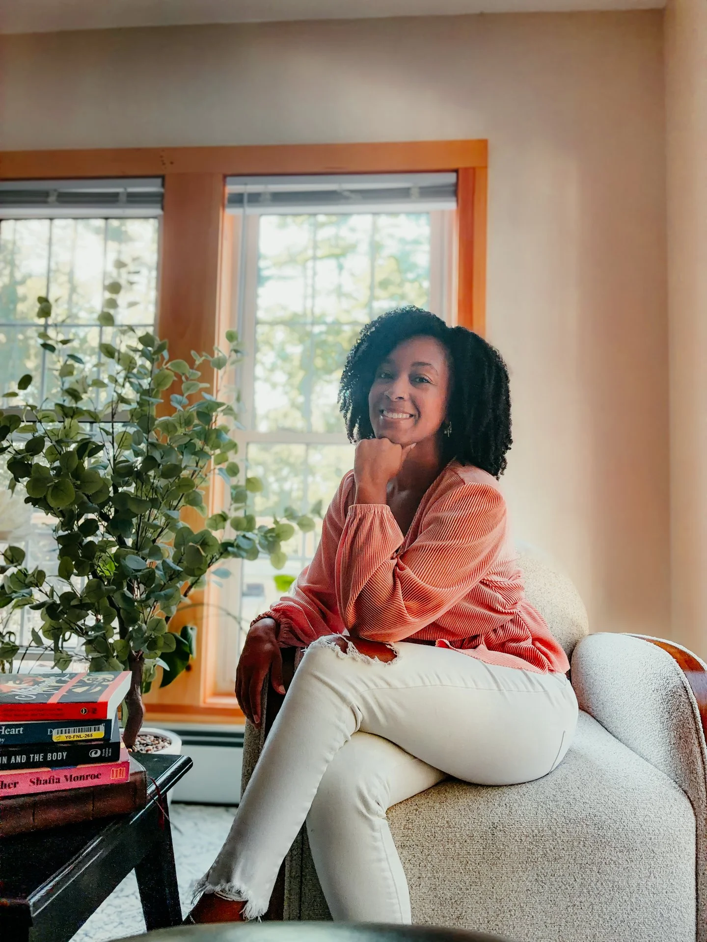 A woman with curly black hair sitting on a beige couch in a living room, smiling and resting her chin on her hand. She is wearing a coral-colored, long-sleeve top and white ripped jeans. There is a large green plant beside her, a stack of books on a black table, and large windows with trees outside in the background.