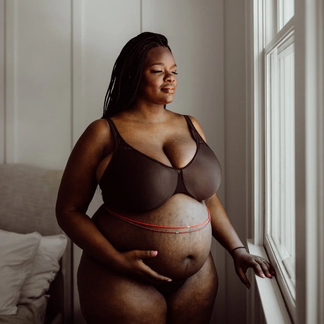 A pregnant woman stands by a window, gently touching her belly, wearing a sheer black bra and black underwear.