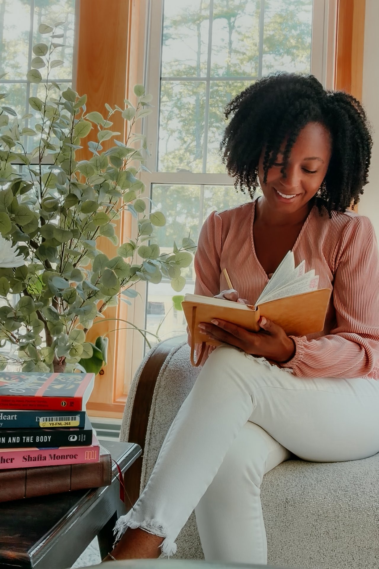 A woman with curly black hair wearing a pink striped blouse and white pants sitting on a beige armchair near a large window, smiling while reading a notebook. There is a tall green plant and a stack of books on a nearby table.