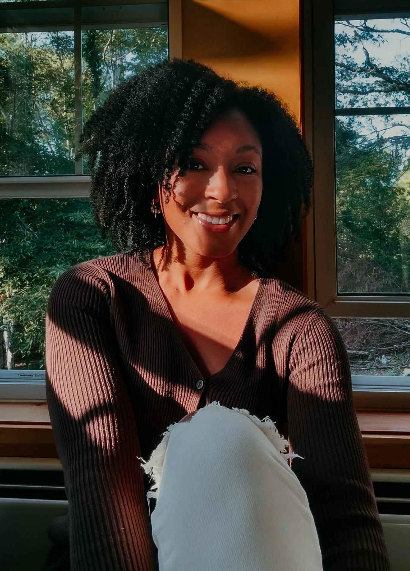 A woman with curly black hair smiling, seated by a large window showing green trees outside, wearing a brown ribbed long-sleeve shirt and earrings, with sunlight illuminating her face.
