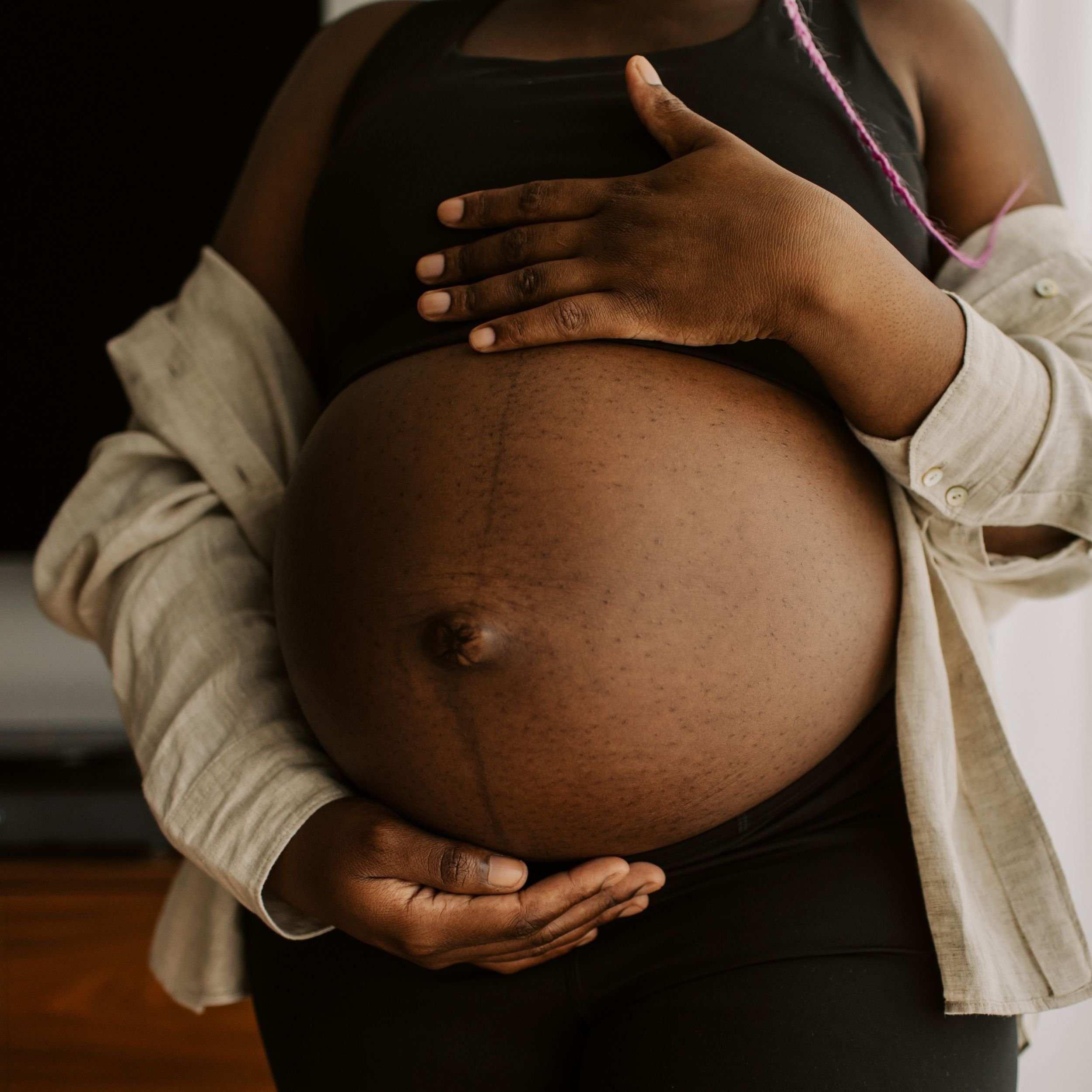 A person with dark skin tone holding their pregnant belly with one hand, wearing a black top and a beige or light-colored cardigan. The focus is on the belly and hands, with the person's face not visible.