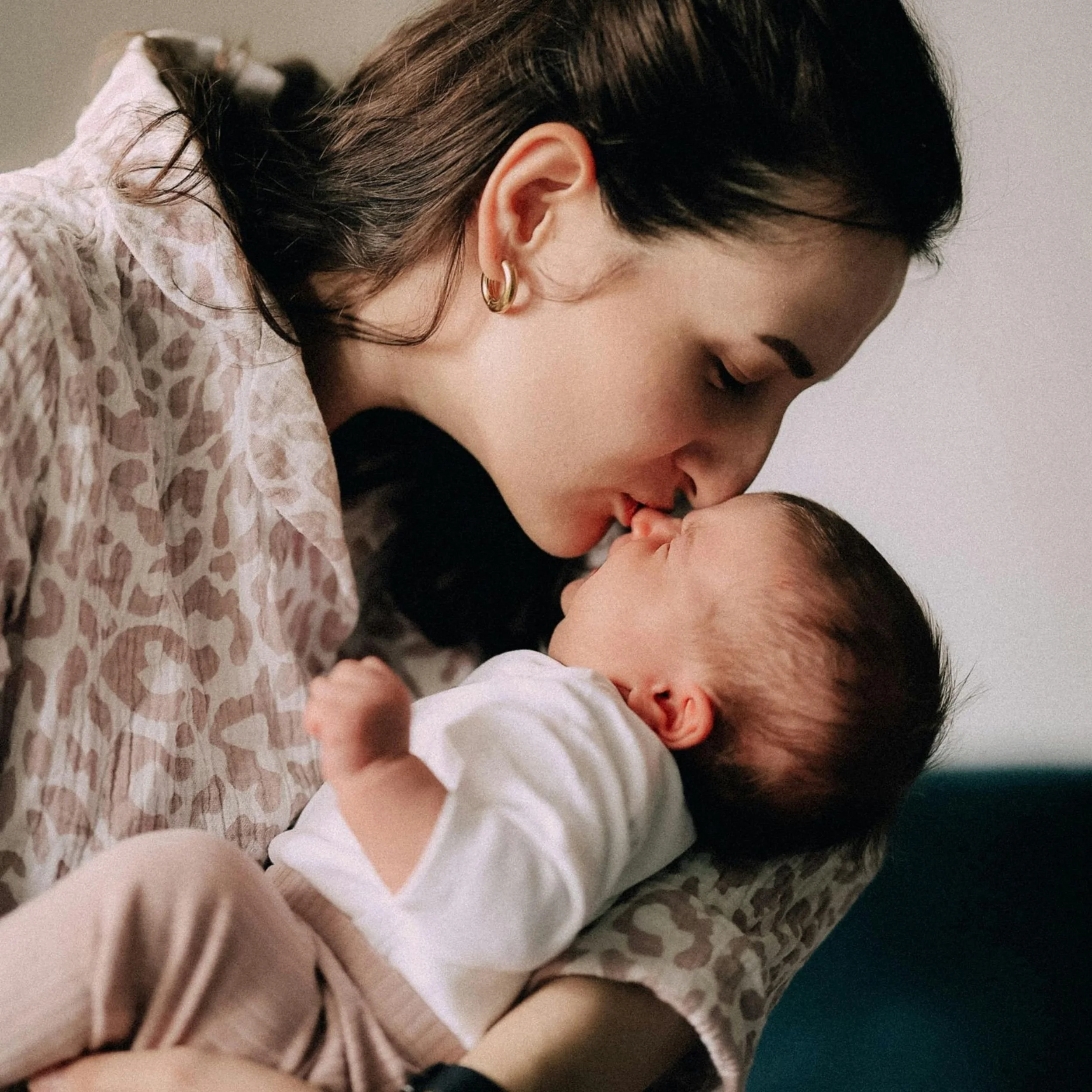 A woman with short dark hair, wearing earrings, gently kisses a baby on the nose. The woman is wearing a patterned shirt and is leaning close to the baby's face.