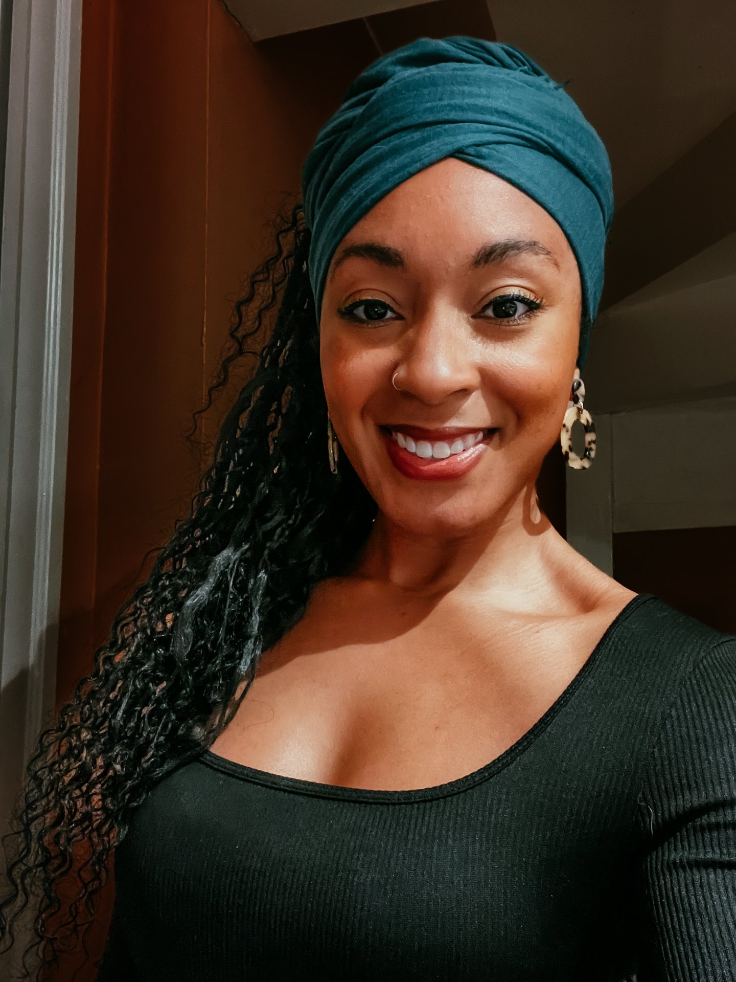 A smiling woman with dark, curly hair wrapped in a teal headscarf, wearing a black top with a rounded neckline, in a room with wooden and gray walls.