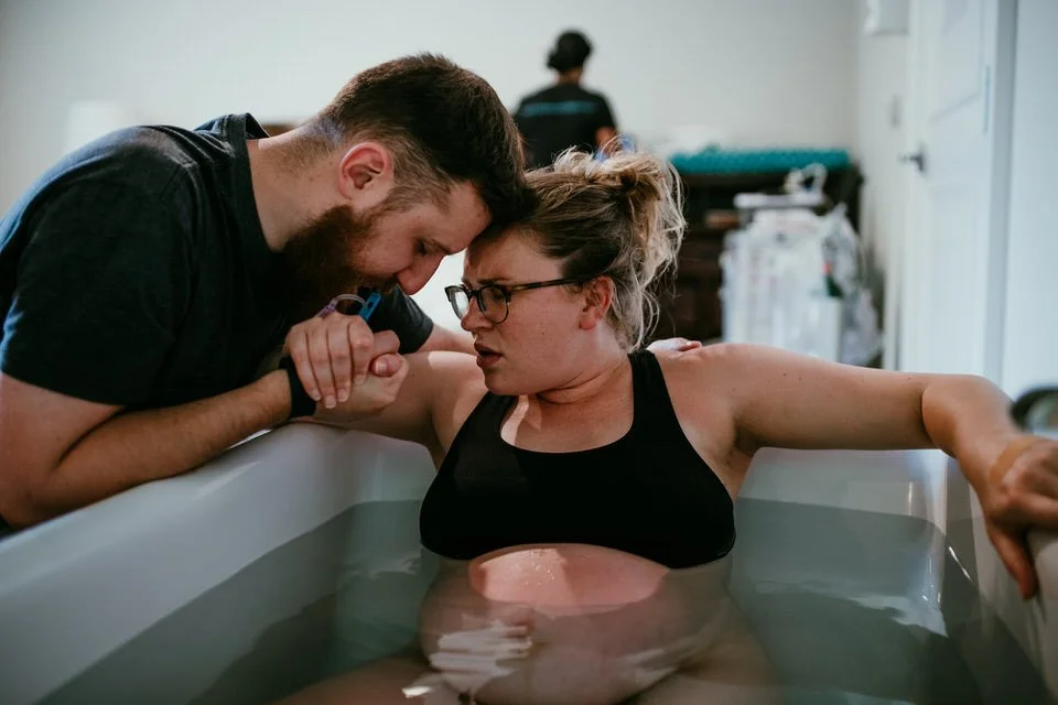 A woman with glasses and a bun in a black tank top is giving birth in a bathtub, supported by a man with a beard who is holding her hand and providing comfort.