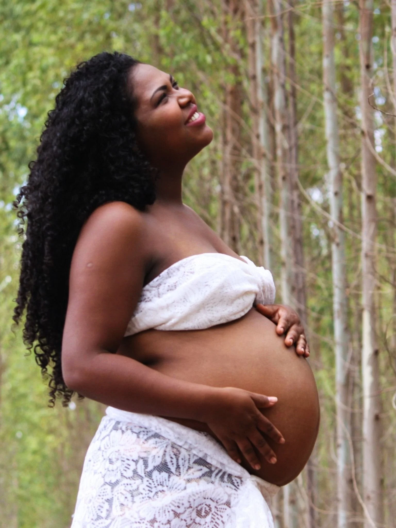 A pregnant woman with long curly black hair and dark skin in a white off-shoulder dress, standing outdoors among tall trees with green leaves, holding her belly with both hands and smiling.