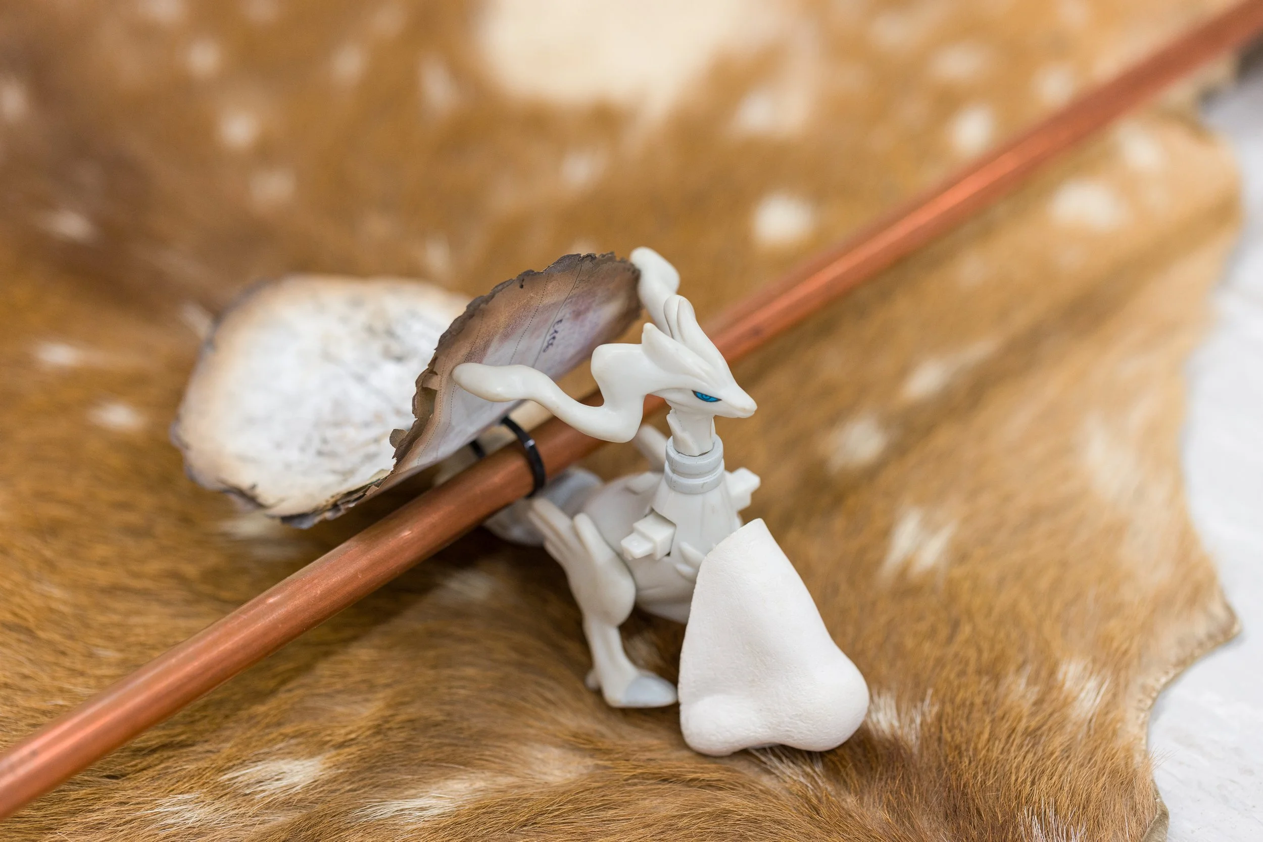 Ein Drachen-Logo mit weißen Gliedmaßen und blauen Augen sitzt auf einer weißen Kugel, neben einem großen Muschelschalenstück, alles auf einem braunen Tierfell. Es wird eine lange Waffe oder Stab gehalten.