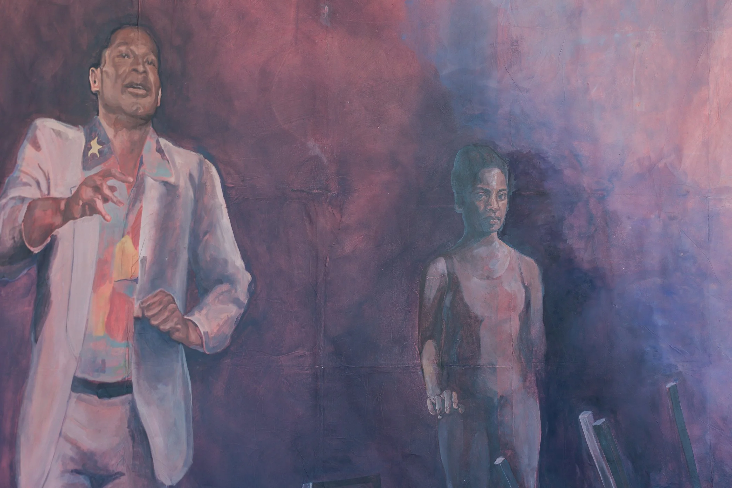 Eine Wandmalerei zeigt eine ältere Frau in einem weißen Kleid mit einem Stern auf der Schulter, die spricht oder erklärt, während eine jüngere Frau in einem hellen Kleid daneben steht, auf einen Gegenstand oder eine Tafel schauend.