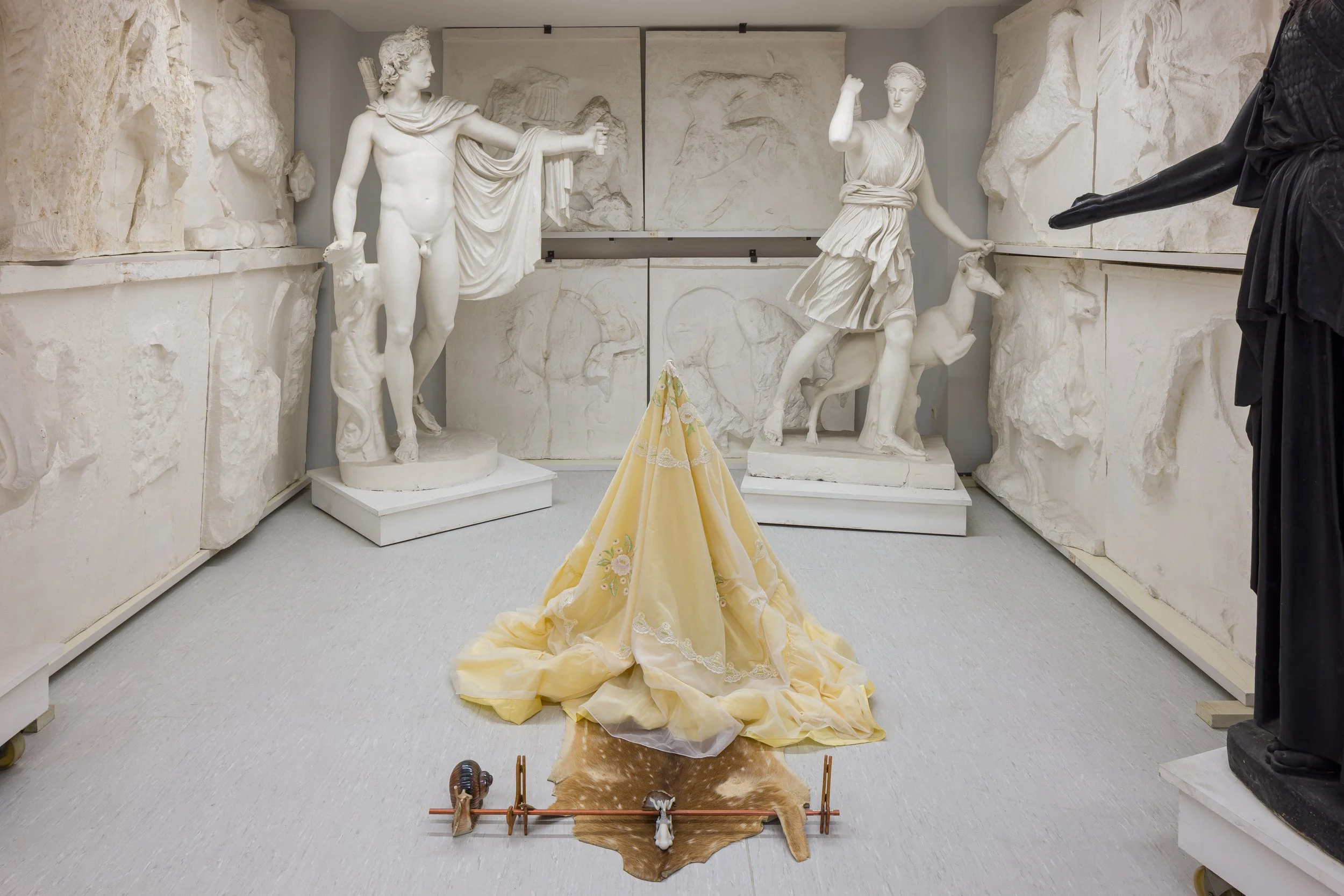 Ein Raum mit antiken weißen Marmorskulpturen und einem gelben Stoff, der wie ein Kleid aussieht, mit einem Tierfell darunter, vermutlich in einem Museum oder einer Sammlung. Die Skulpturen zeigen antike Figuren, einige mit Tieren.