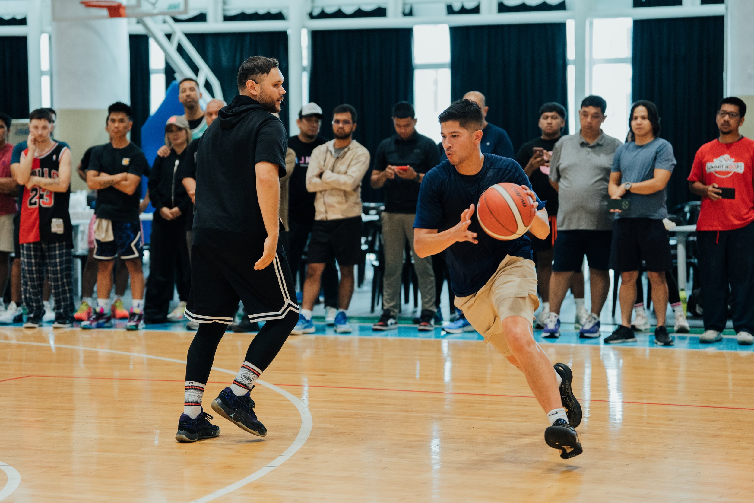 Two men playing basketball on an indoor court, with a group of spectators watching in the background.