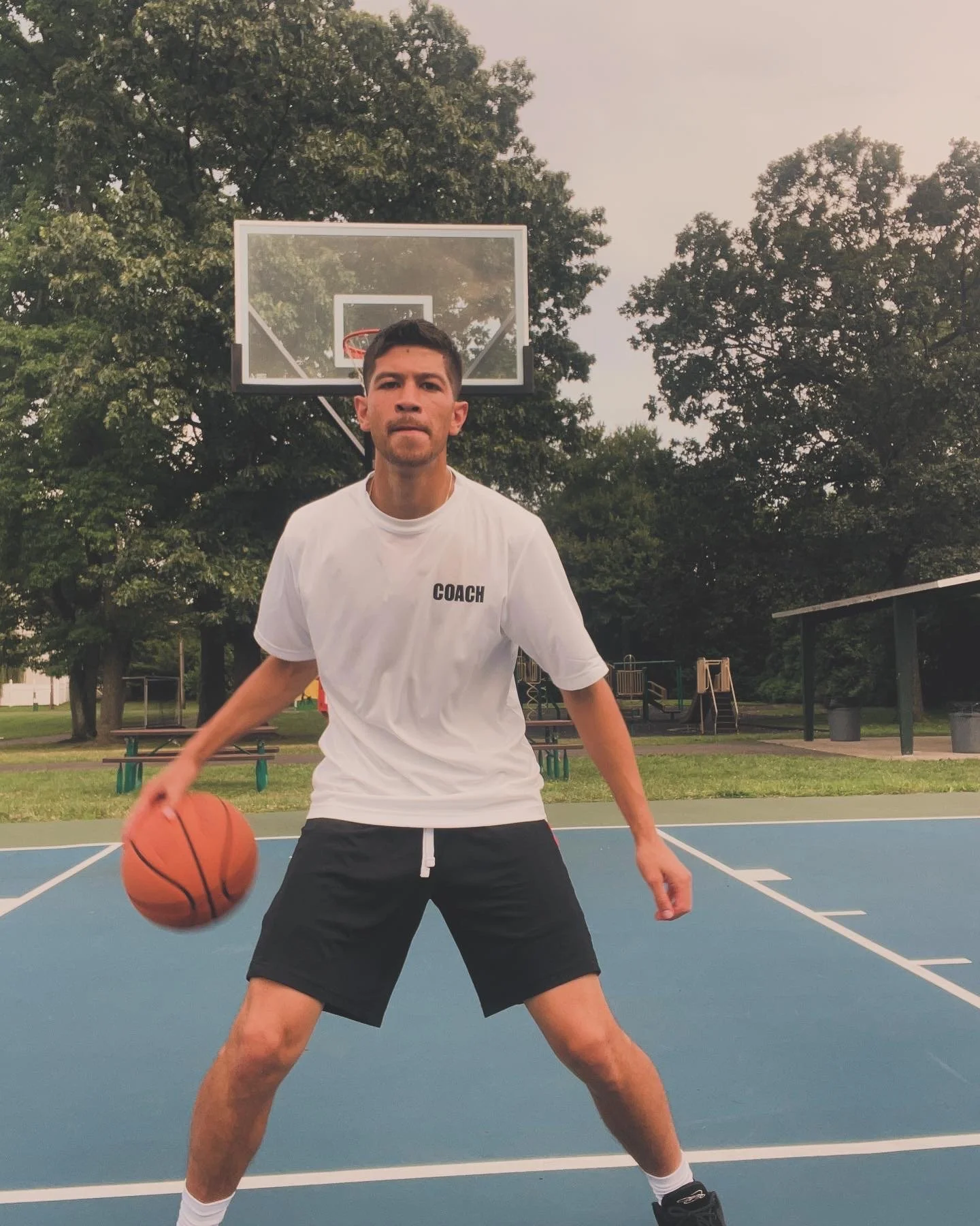 A man wearing a white T-shirt labeled 'COACH' and black shorts, playing basketball on an outdoor court surrounded by trees.