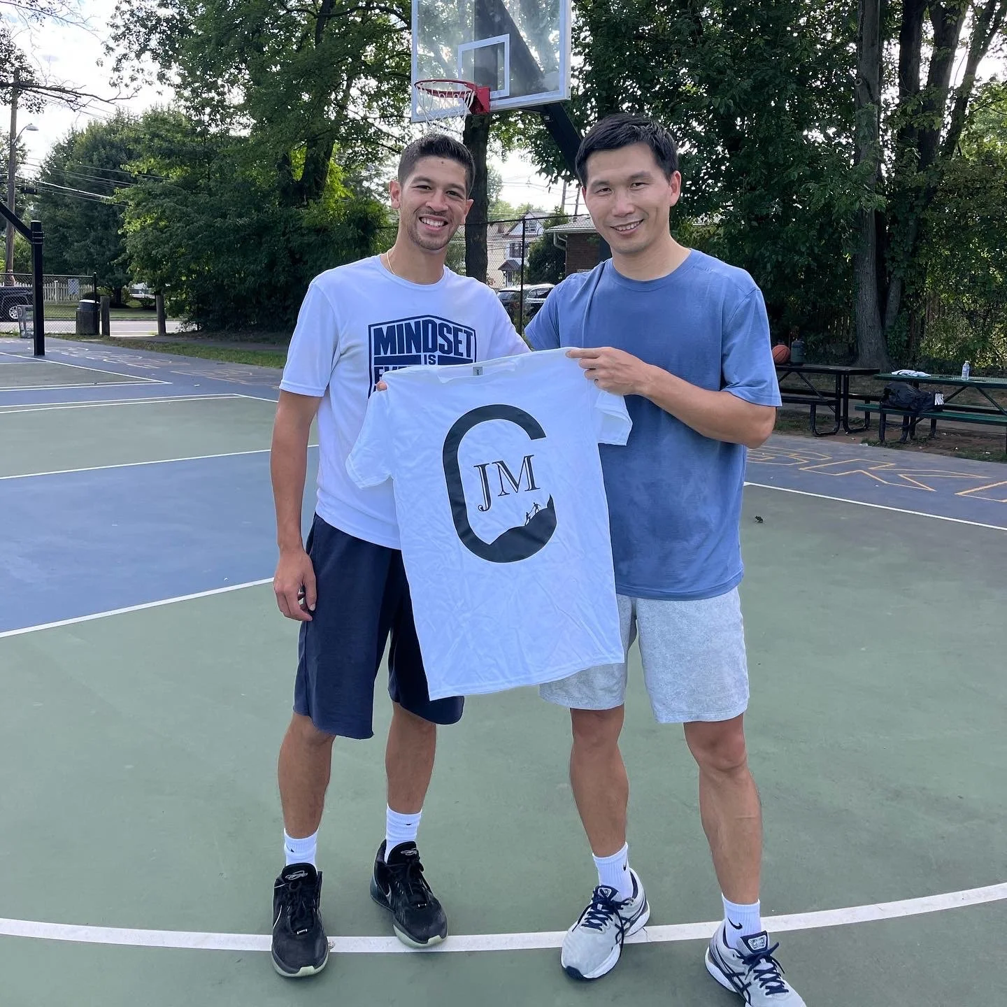 Two men standing on a basketball court, smiling, holding a white T-shirt with a logo and initials, with trees and a basketball hoop in the background.