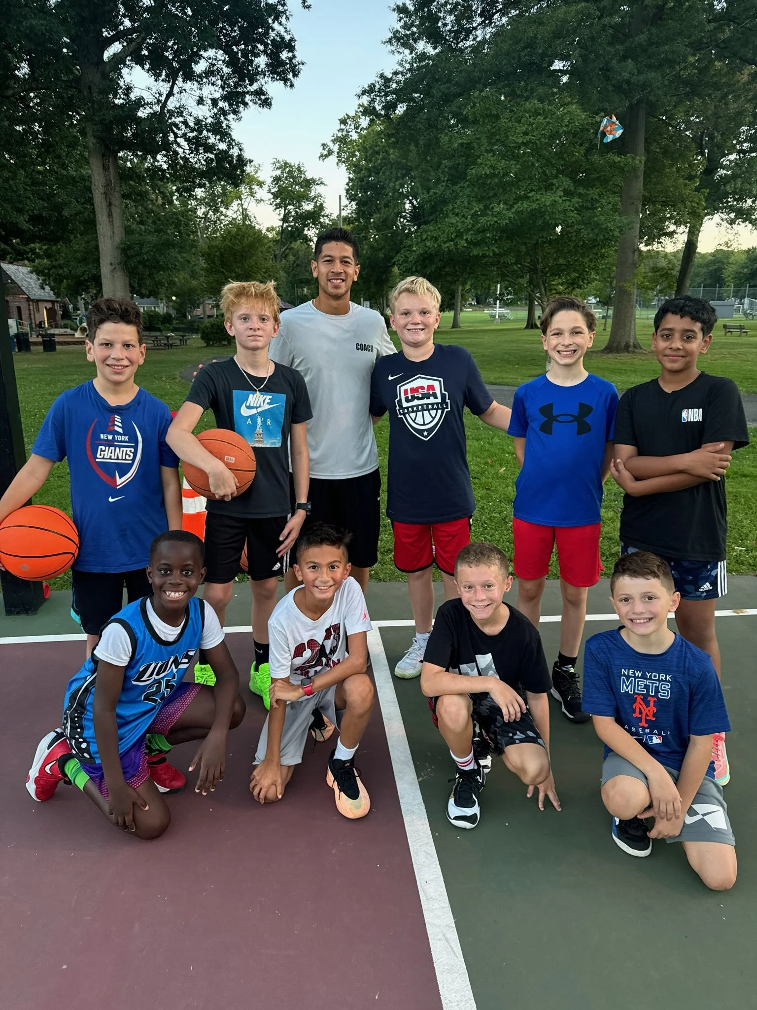 A group of young boys and their coach posing on a basketball court at sunset. The boys are holding basketballs and wearing sportswear, some with team logos from New York Giants, Nike, USA Basketball, Under Armour, NBA, and Mets. They are smiling and 