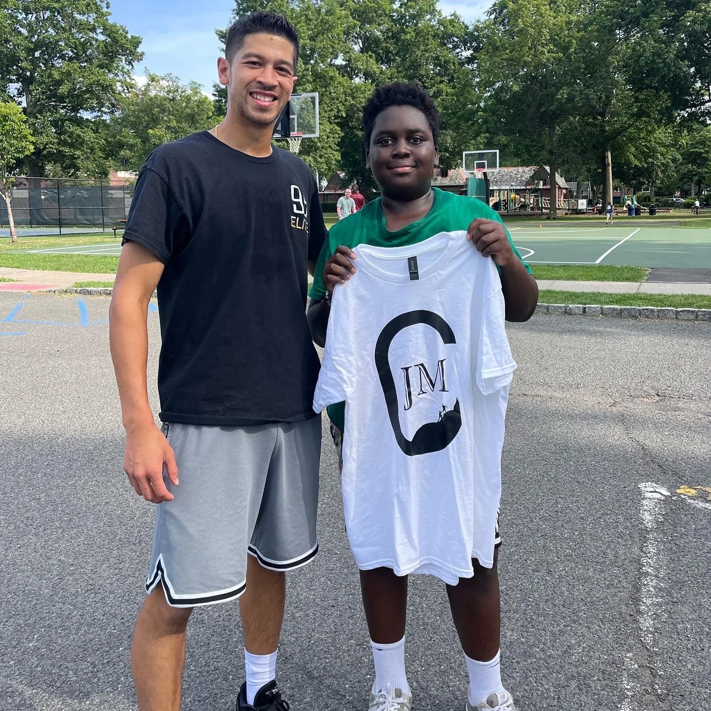 A young boy holding a white t-shirt with a mustache and glasses graphic, standing next to a smiling young man on a basketball court with trees and playground equipment in the background.