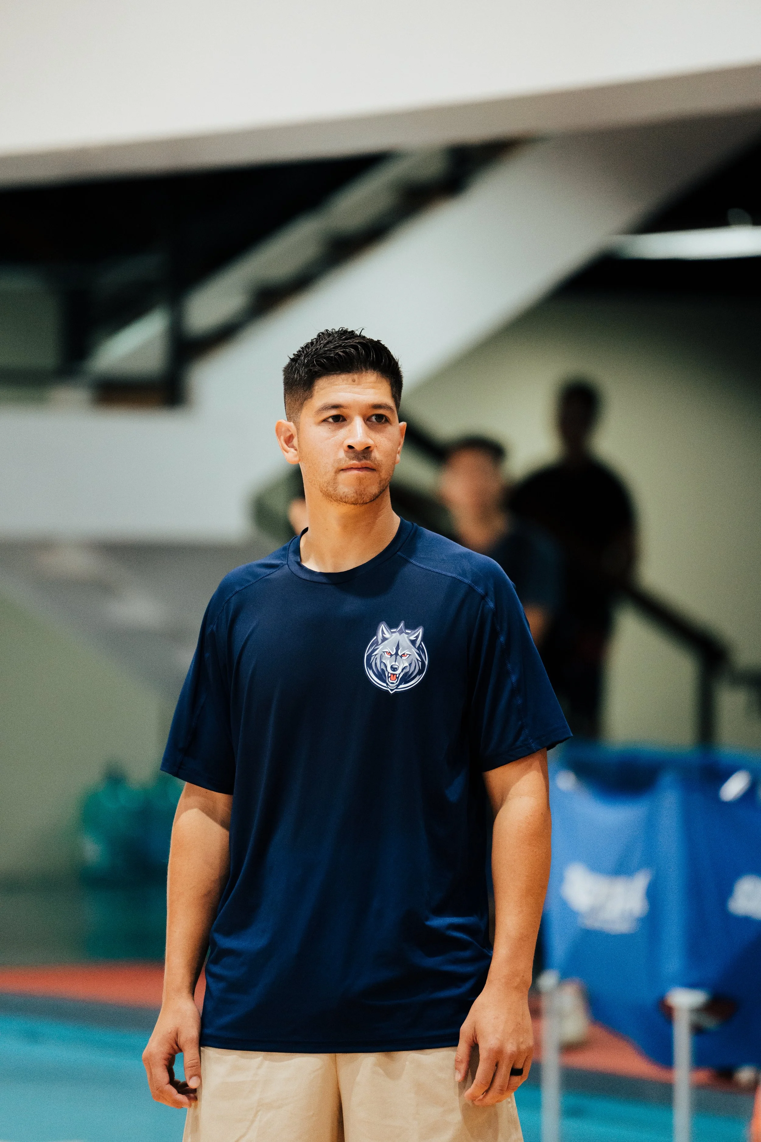 A young man with short black hair wearing a navy blue sports shirt with a wolf logo, standing indoors near a sports court.