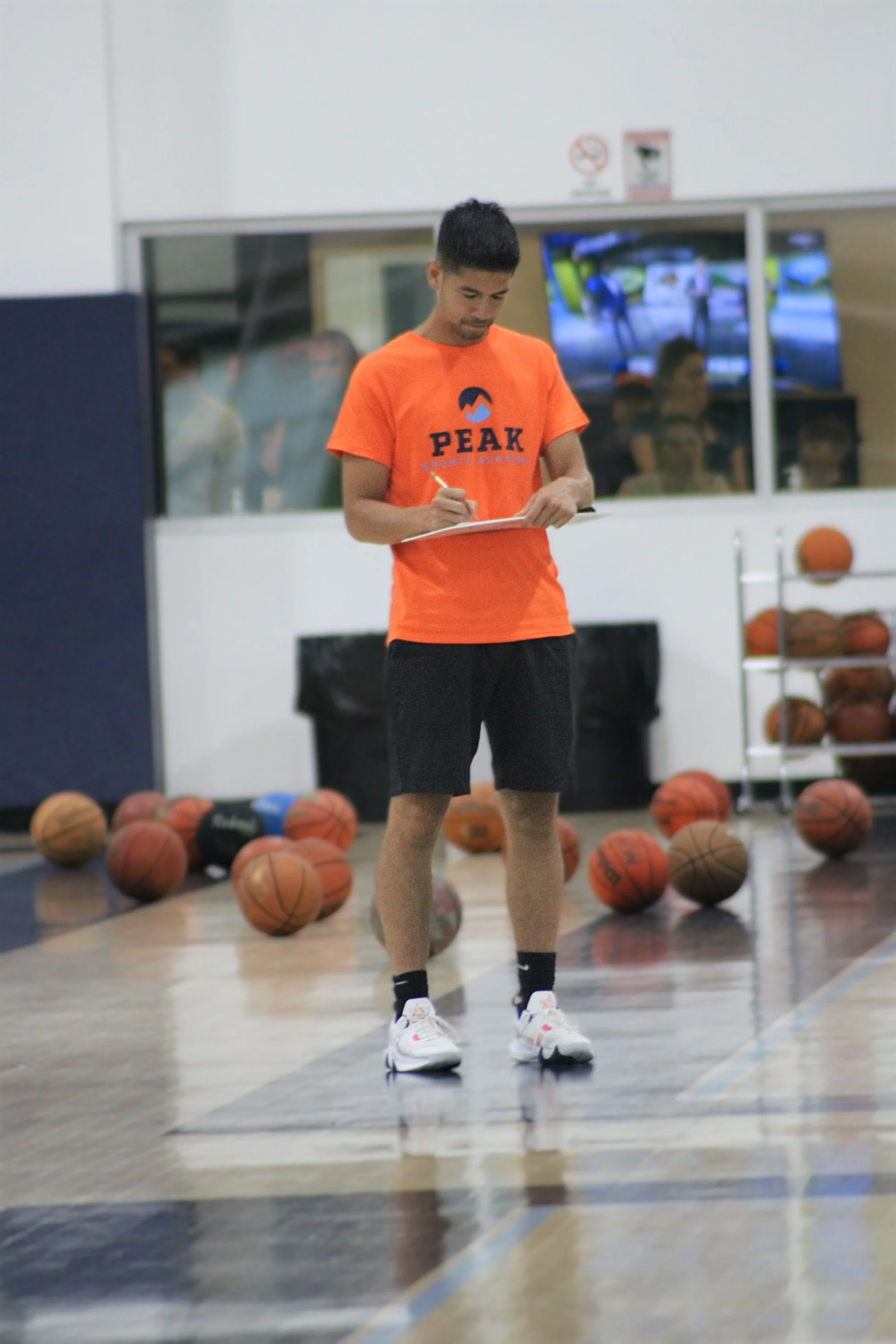 A young male basketball player in an orange shirt with 'PEAK' printed on it, taking notes or writing on a clipboard in a gymnasium with basketballs on the floor behind him.