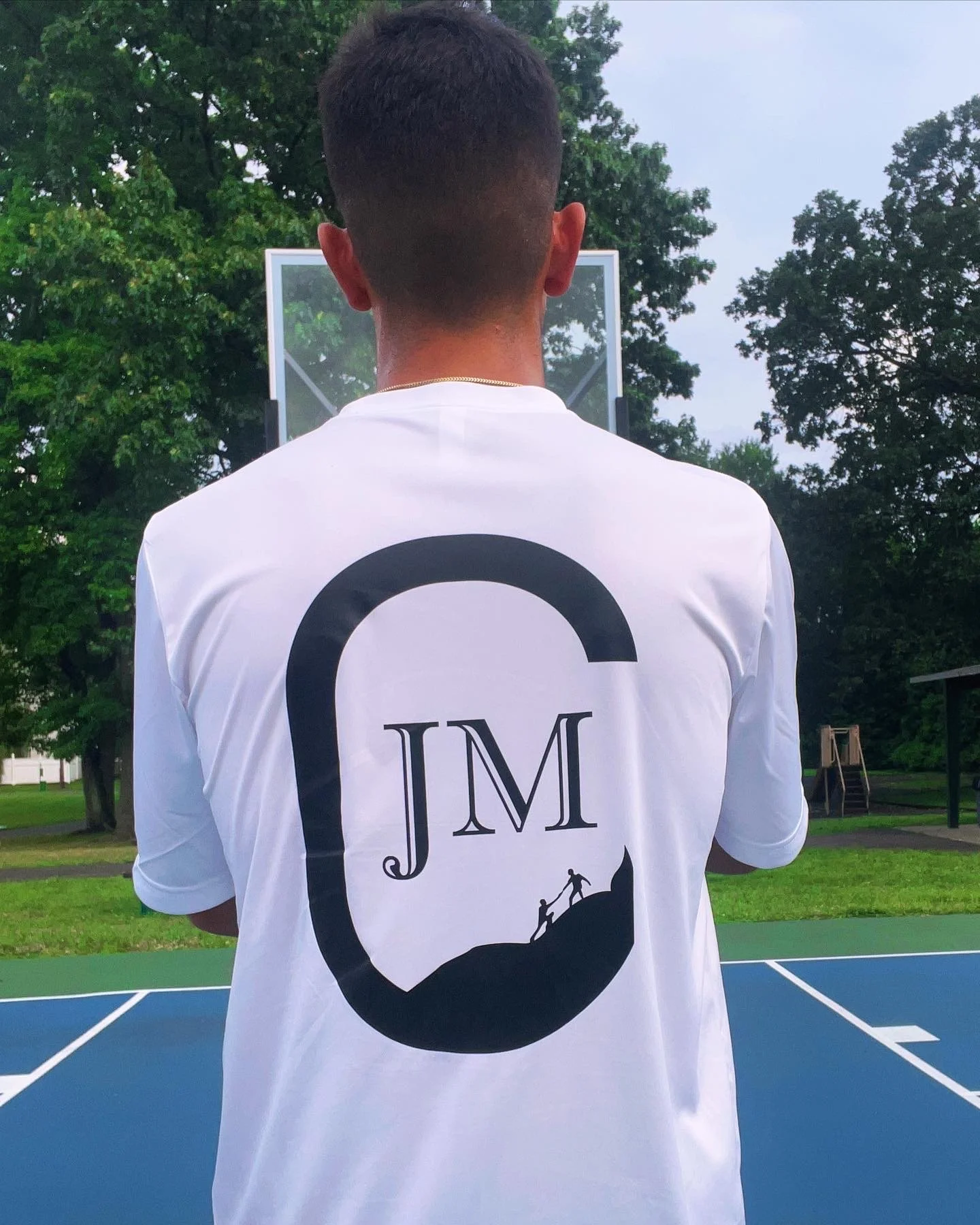 A person is standing on an outdoor basketball court, facing away from the camera, wearing a white t-shirt with a large black graphic and initials 'JM' on the back. The graphic depicts a silhouette of two people climbing a mountain. Trees and a basket