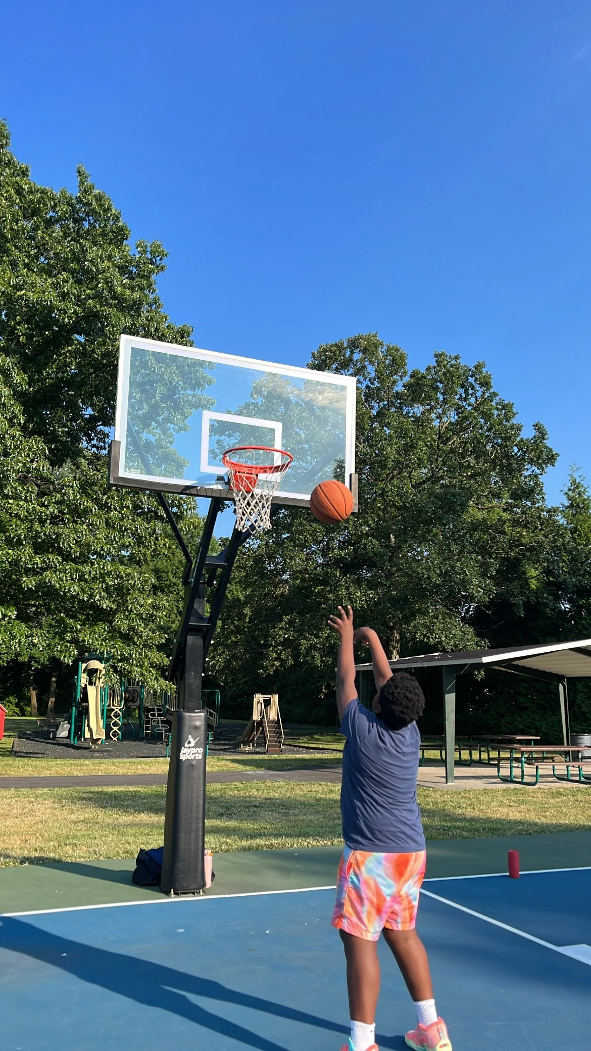 A young boy playing basketball outdoors at a park, shooting a basketball towards the hoop on a sunny day with a clear blue sky and green trees in the background.