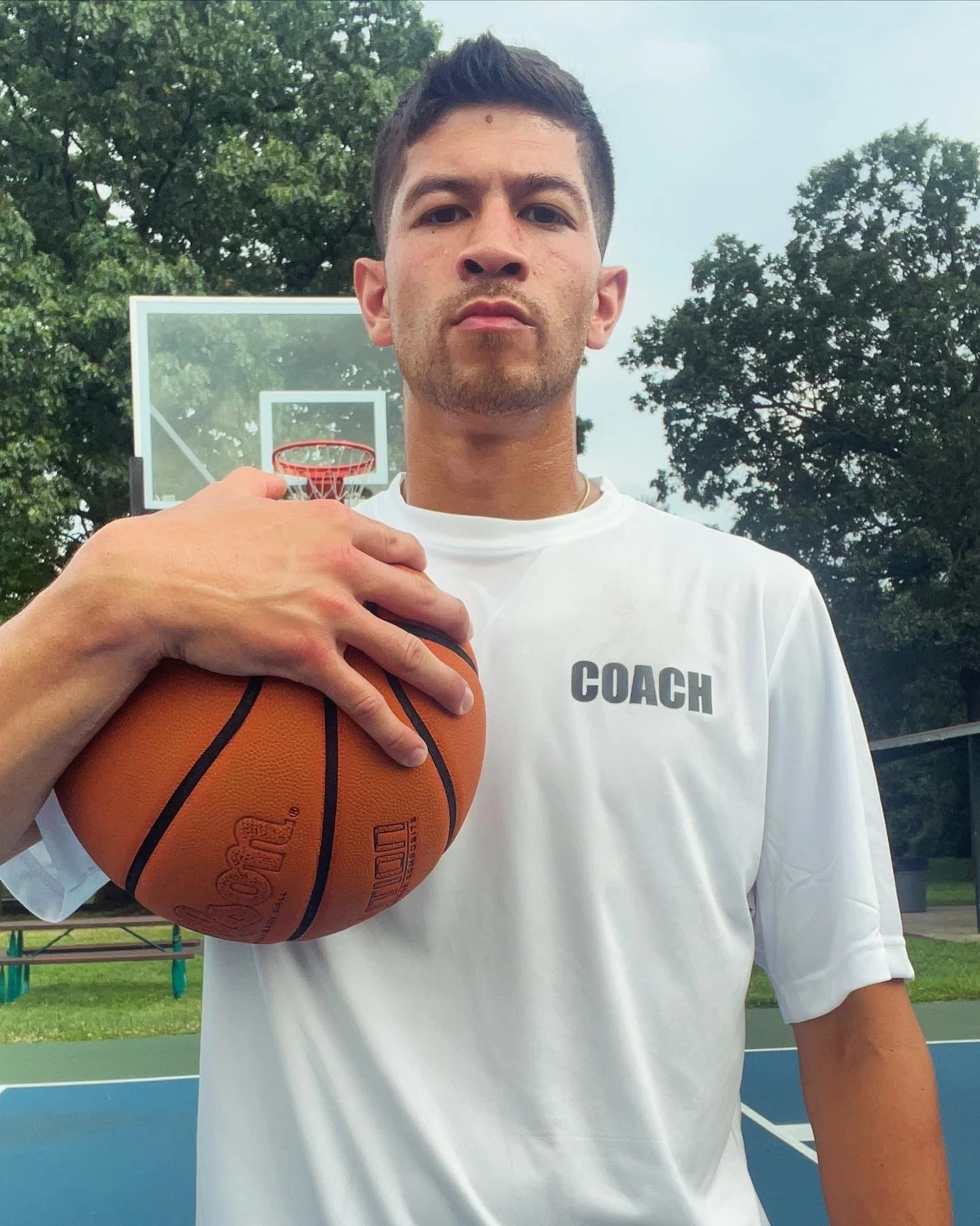 Young man holding a basketball on an outdoor basketball court with a backboard and hoop, wearing a white shirt with 'COACH' printed on it, with trees and a cloudy sky in the background.