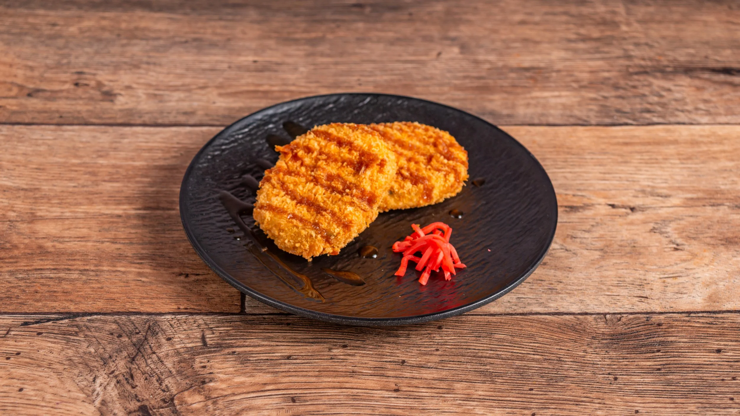 A breaded, fried, pumpkin croquette served on a black plate with a small portion of pickled ginger and a drizzle of sauce, placed on a wooden table.