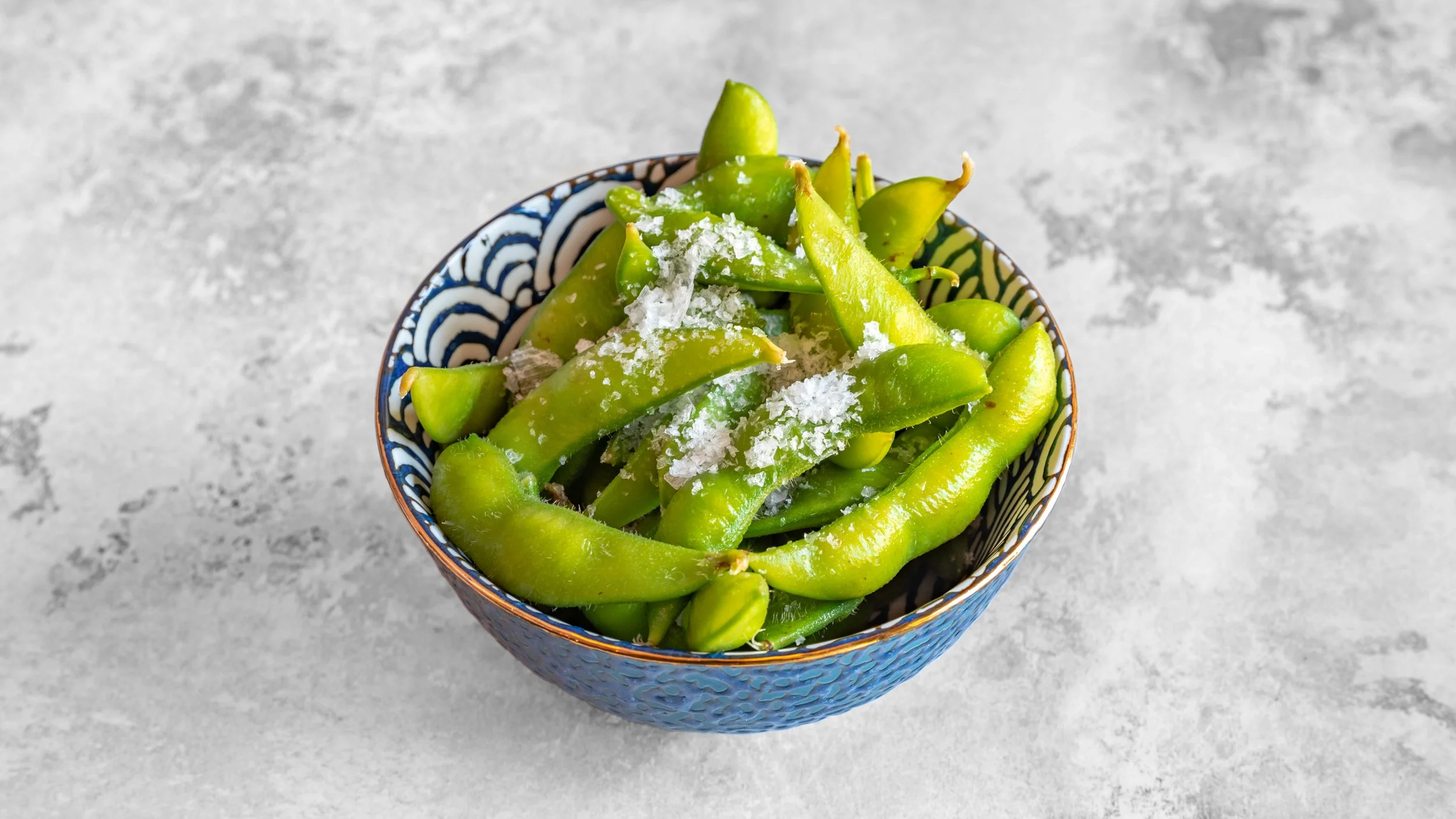 A bowl filled with green soya beans sprinkled with salt, placed on a gray textured surface.