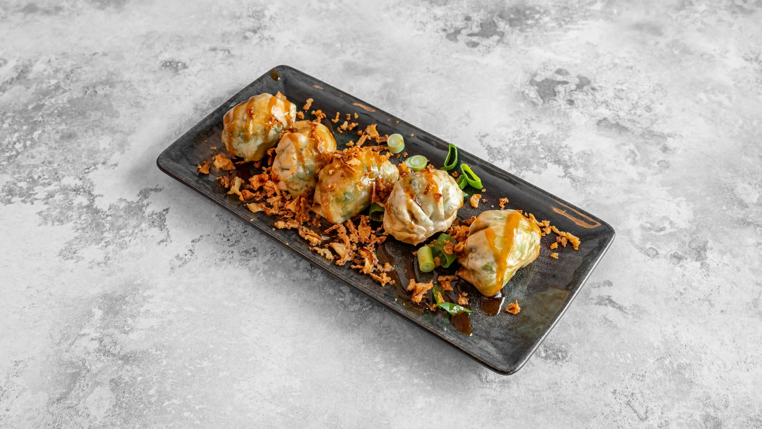 Plate of five Japanese dumplings topped with sauce, garnished with green onions and crispy bits, served on a black rectangular plate.