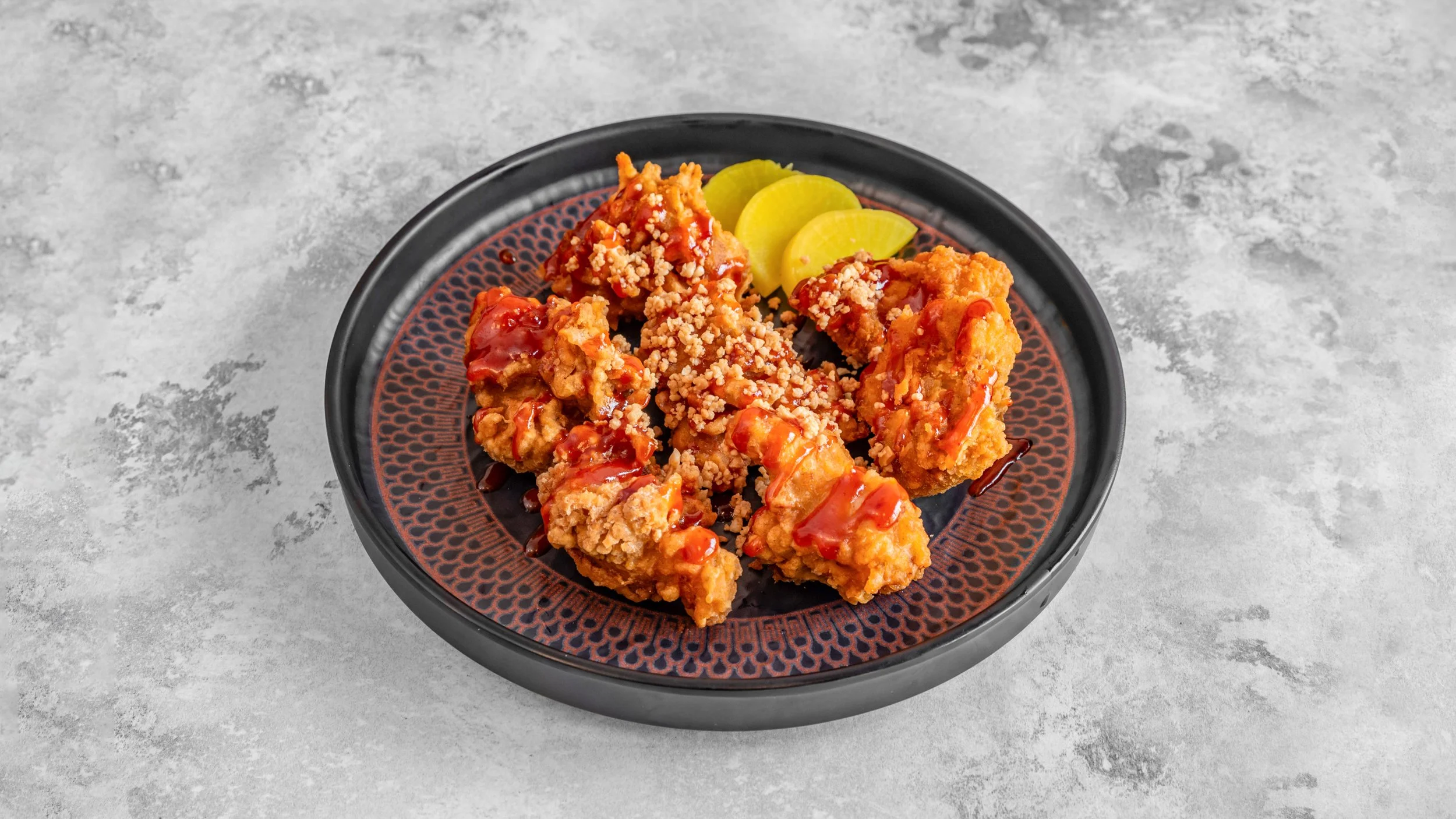 Fried chicken pieces topped with sauce and sprinkled with crushed nuts, served with pickles on a black plate on a concrete surface.