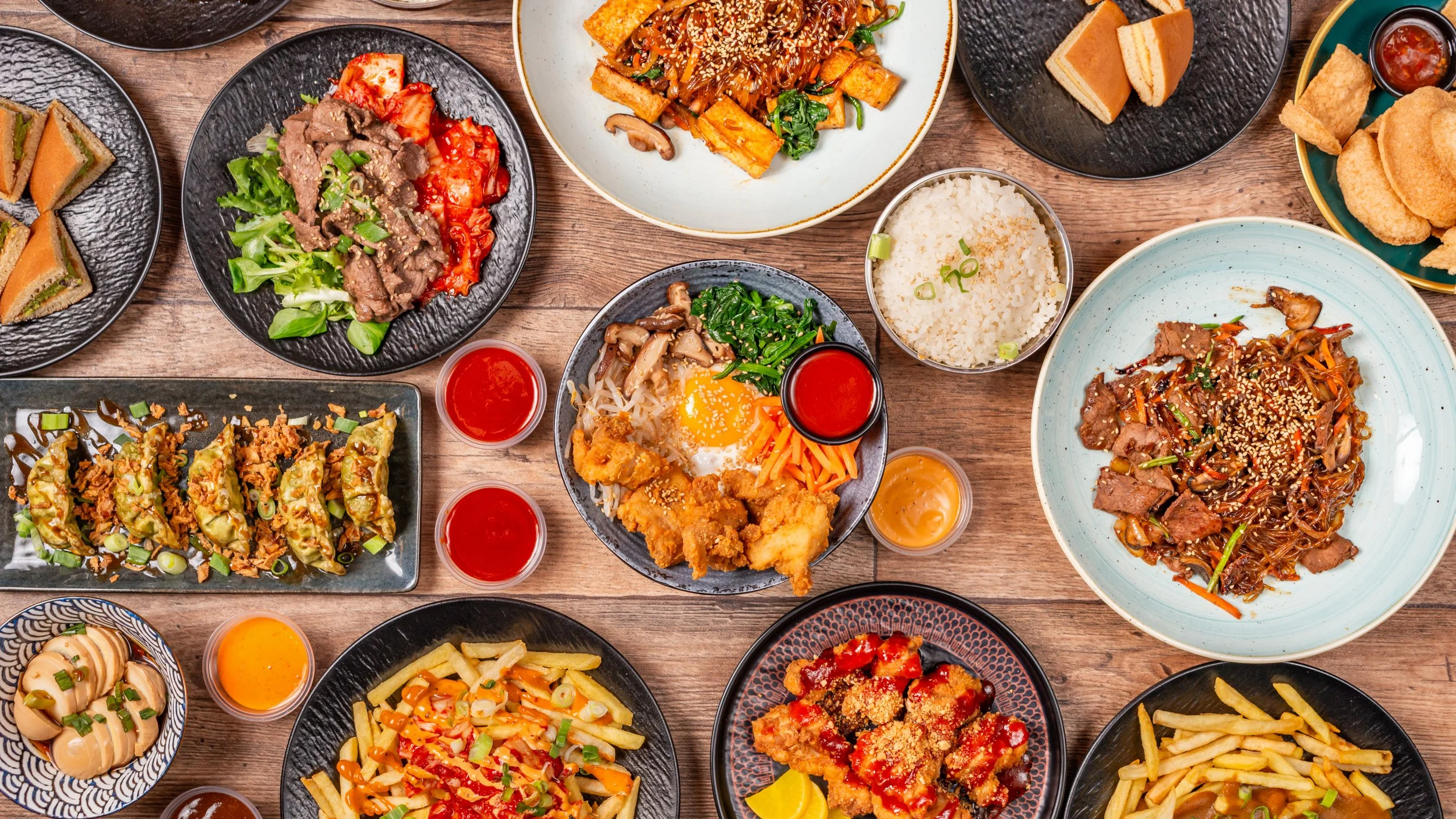 A variety of Asian dishes on black and patterned plates and bowls, including stir-fried noodles, dumplings, fried chicken, rice, vegetables, and small bowls of sauces, all spread out on a wooden table.