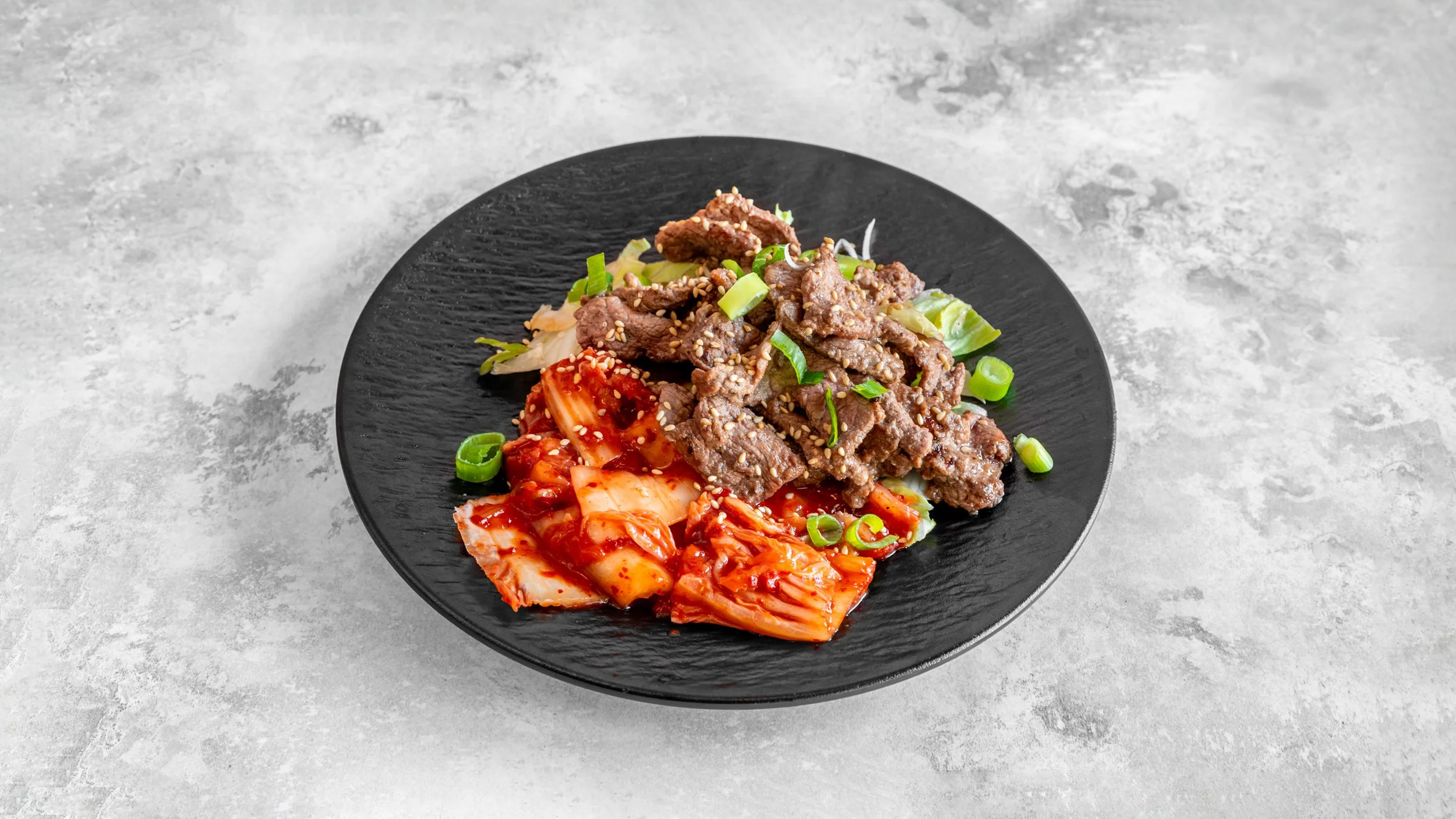Black plate with cooked beef slices topped with chopped green onions and sesame seeds, accompanied by kimchi and a side of spicy red sauce.