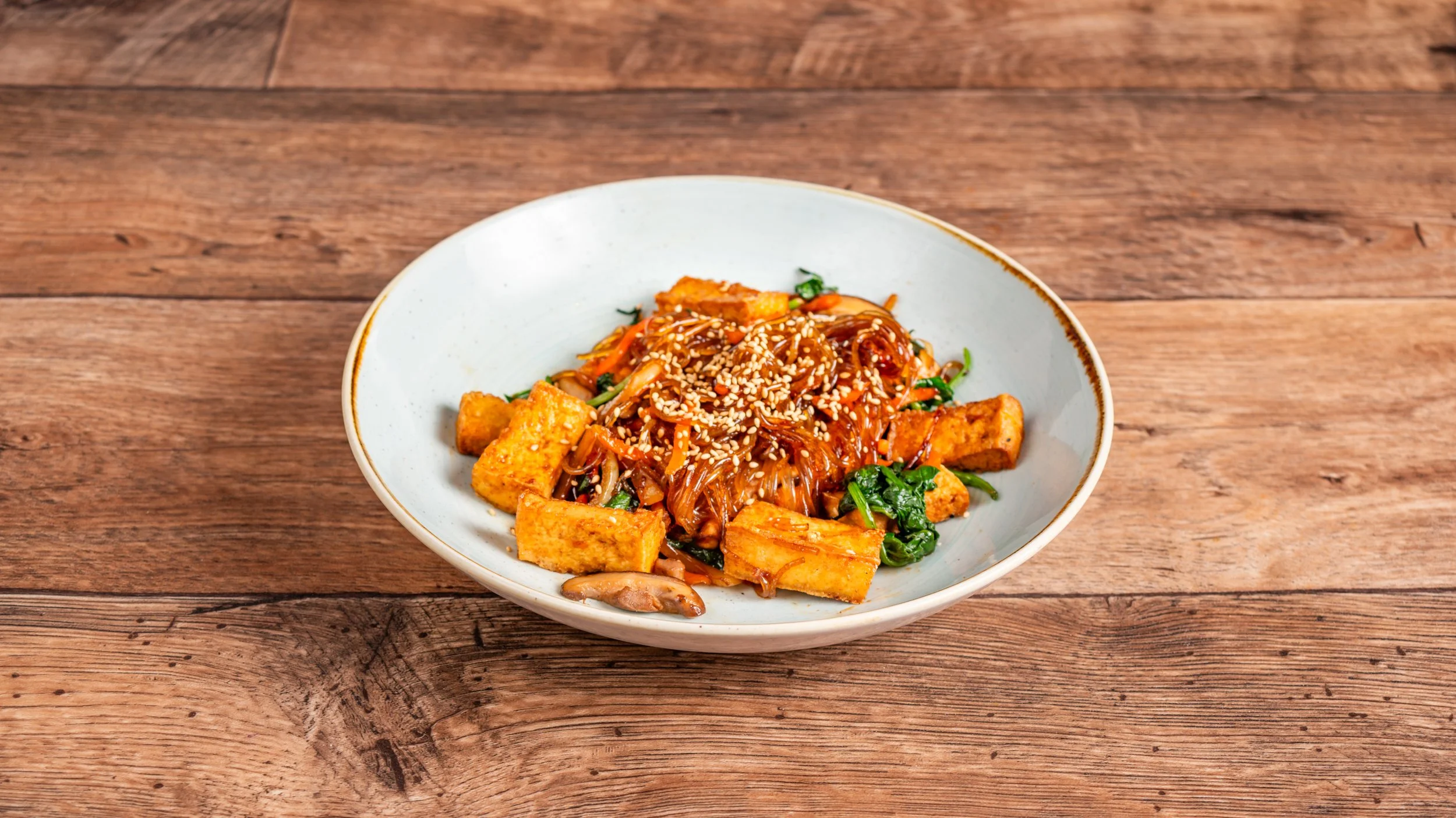 A bowl of Asian stir-fried noodles with tofu, vegetables, and sesame seeds on a wooden table.