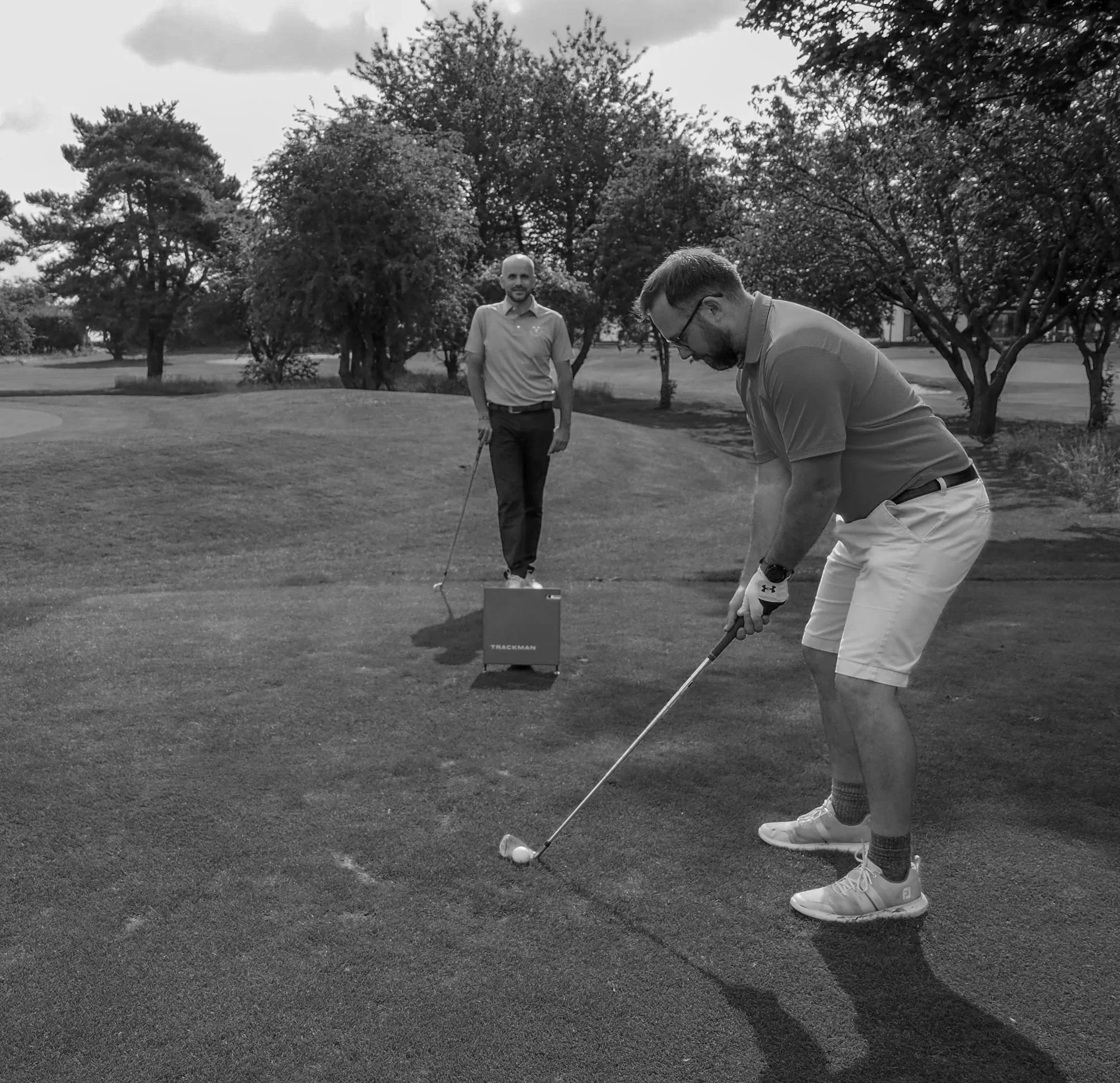 Two men playing golf on a lush green course, with trees and cloudy sky in the background. One man is preparing to hit the golf ball while the other watches, standing behind an orange TrackMan launch monitor.