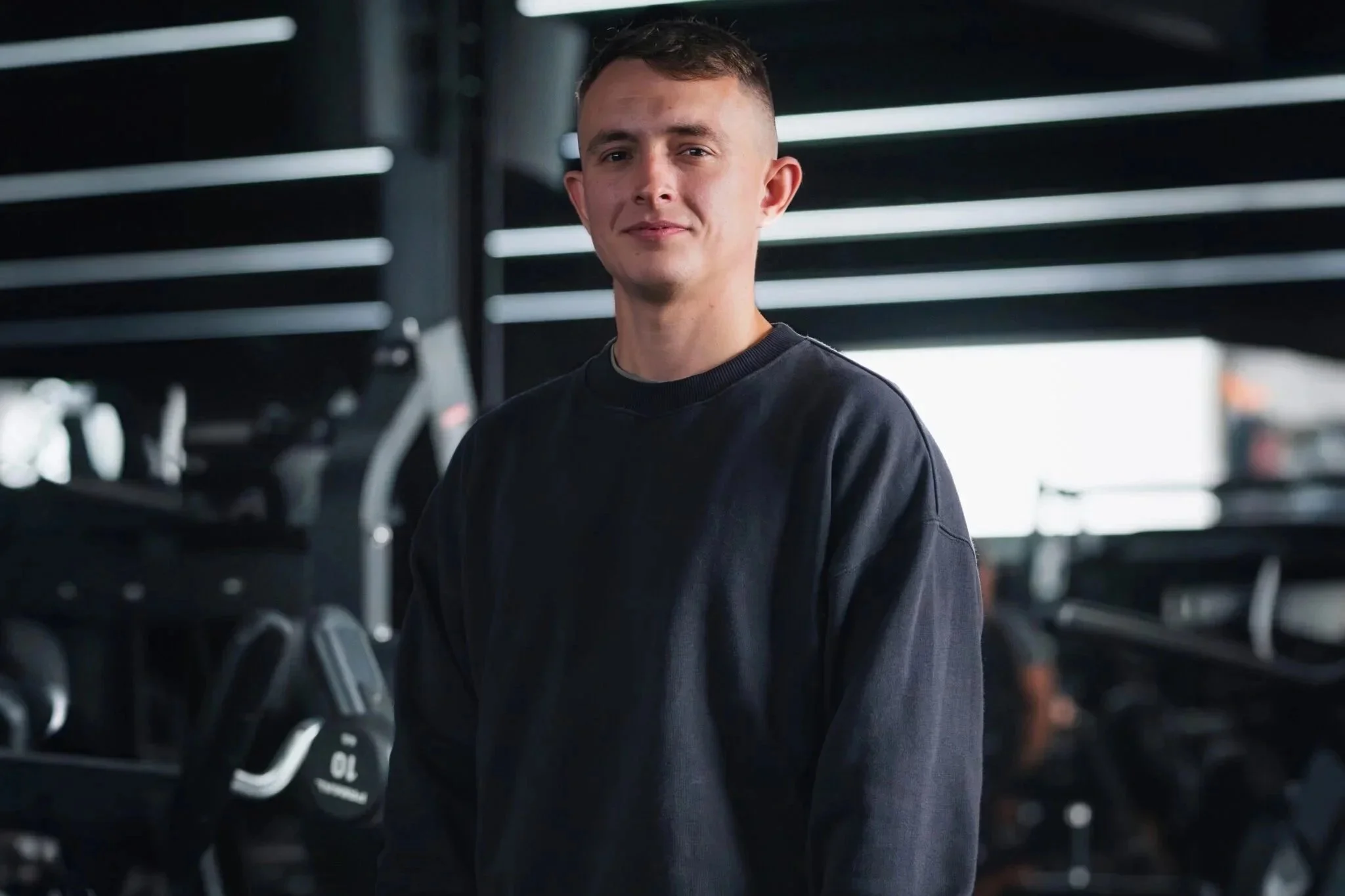 A young man with short brown hair wearing a black sweatshirt standing in a gym with workout equipment in the background.