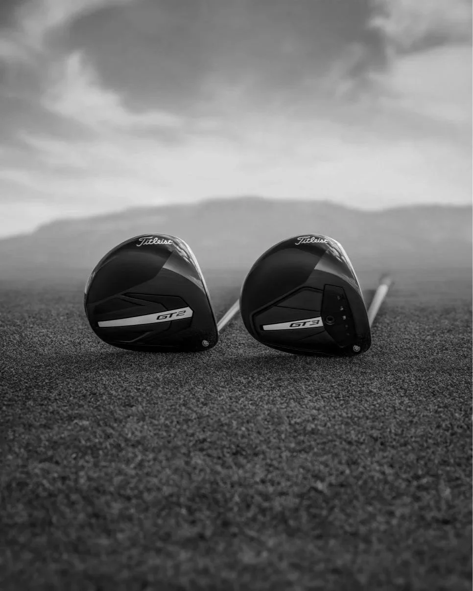 Two golf drivers on grass with mountains and cloudy sky in the background.