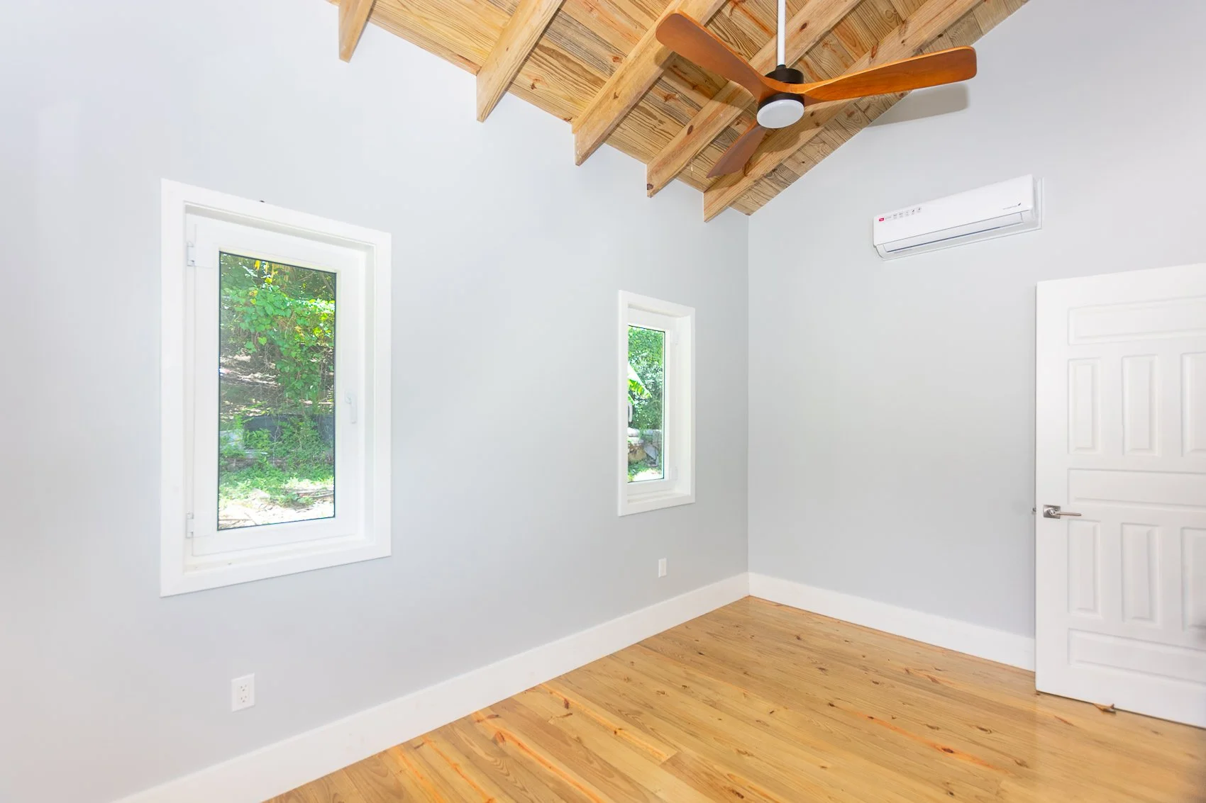 Empty room with two windows, light gray walls, wooden floor, ceiling fan, and air conditioning unit.