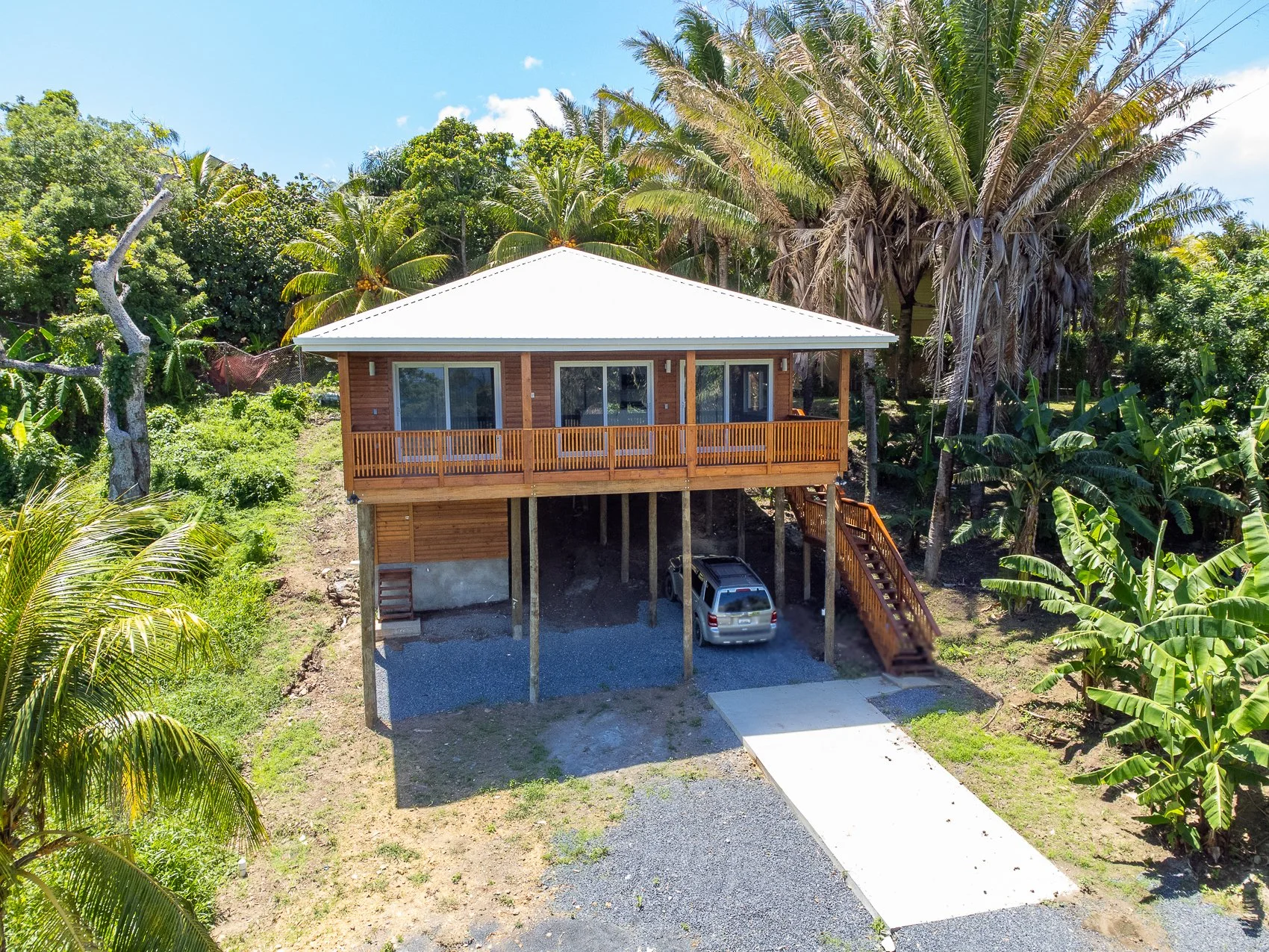 A raised house with a white metal roof on wooden stilts, surrounded by palm trees and lush tropical vegetation, with a driveway and a parked car underneath.