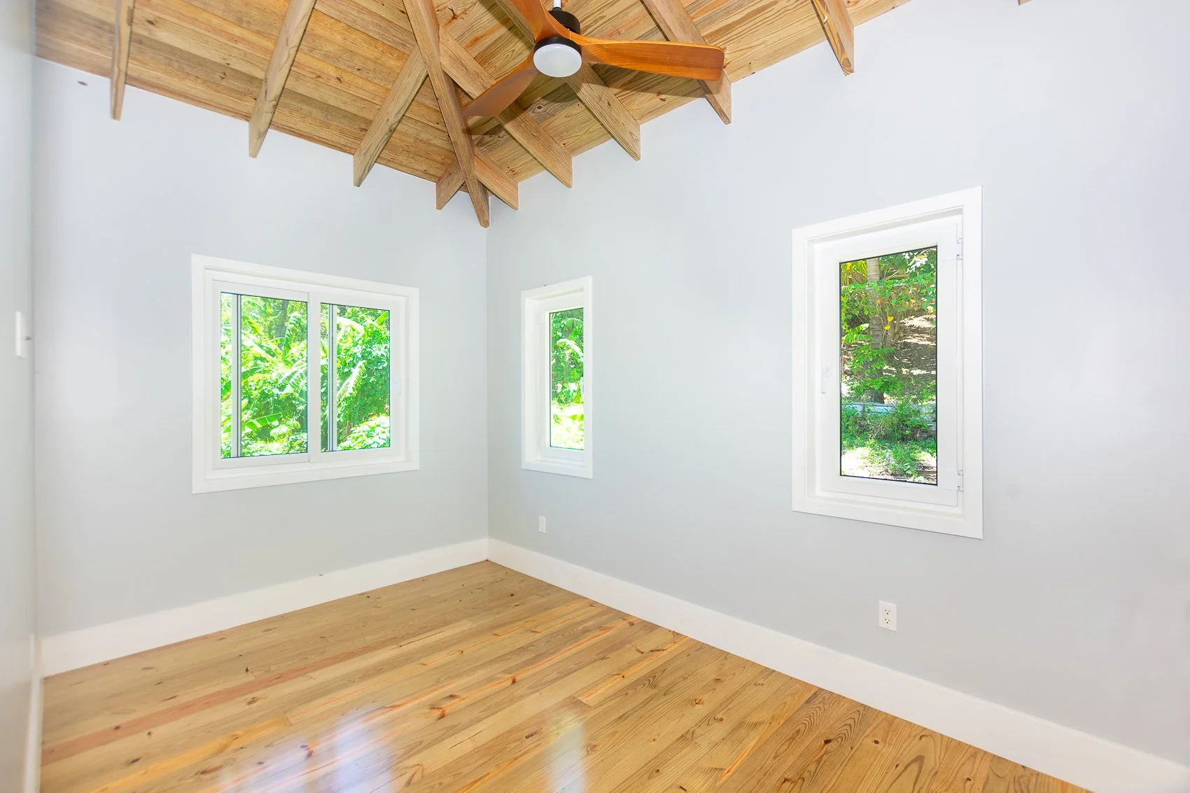 Empty room with light gray walls, wooden floor, three windows showing greenery outside, a ceiling fan, and exposed wooden beams on the ceiling.